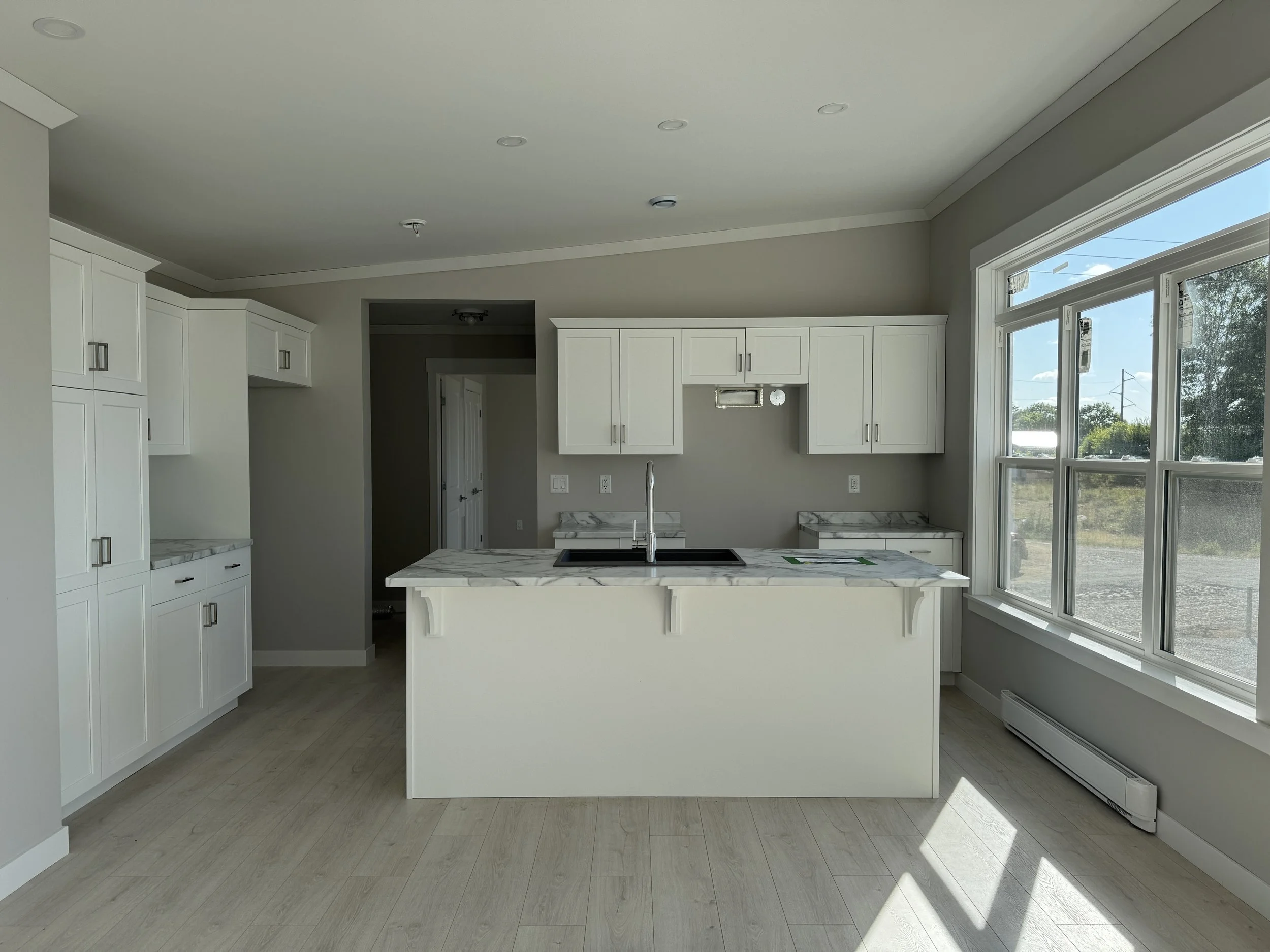 A modern single-story house with black and gray siding, multiple windows, a wooden deck with stairs, a pathway, and landscaped bushes and flowers in front.