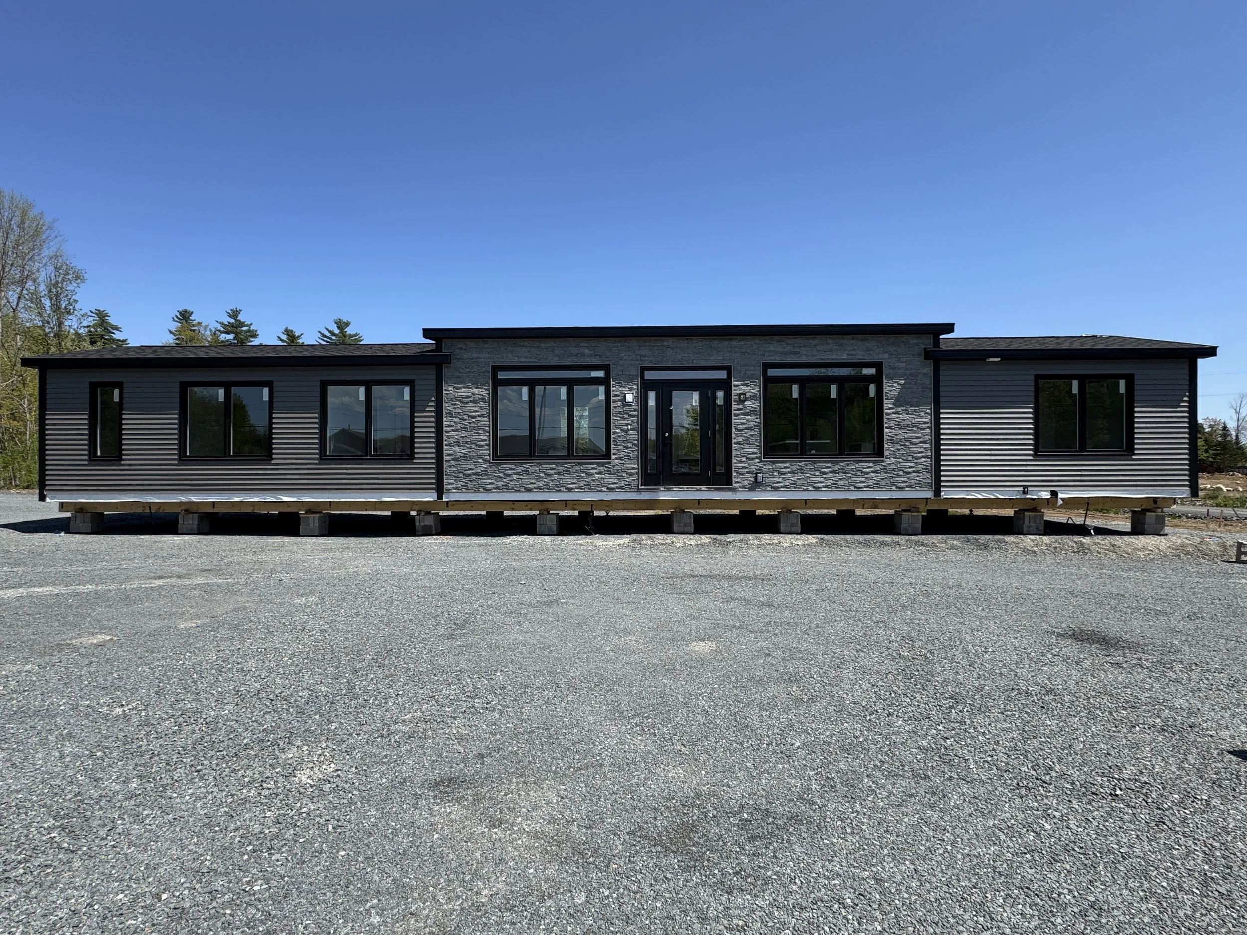 Modern manufactured house with black framing, siding, and stone accents, situated on a gravel lot with a clear blue sky background.