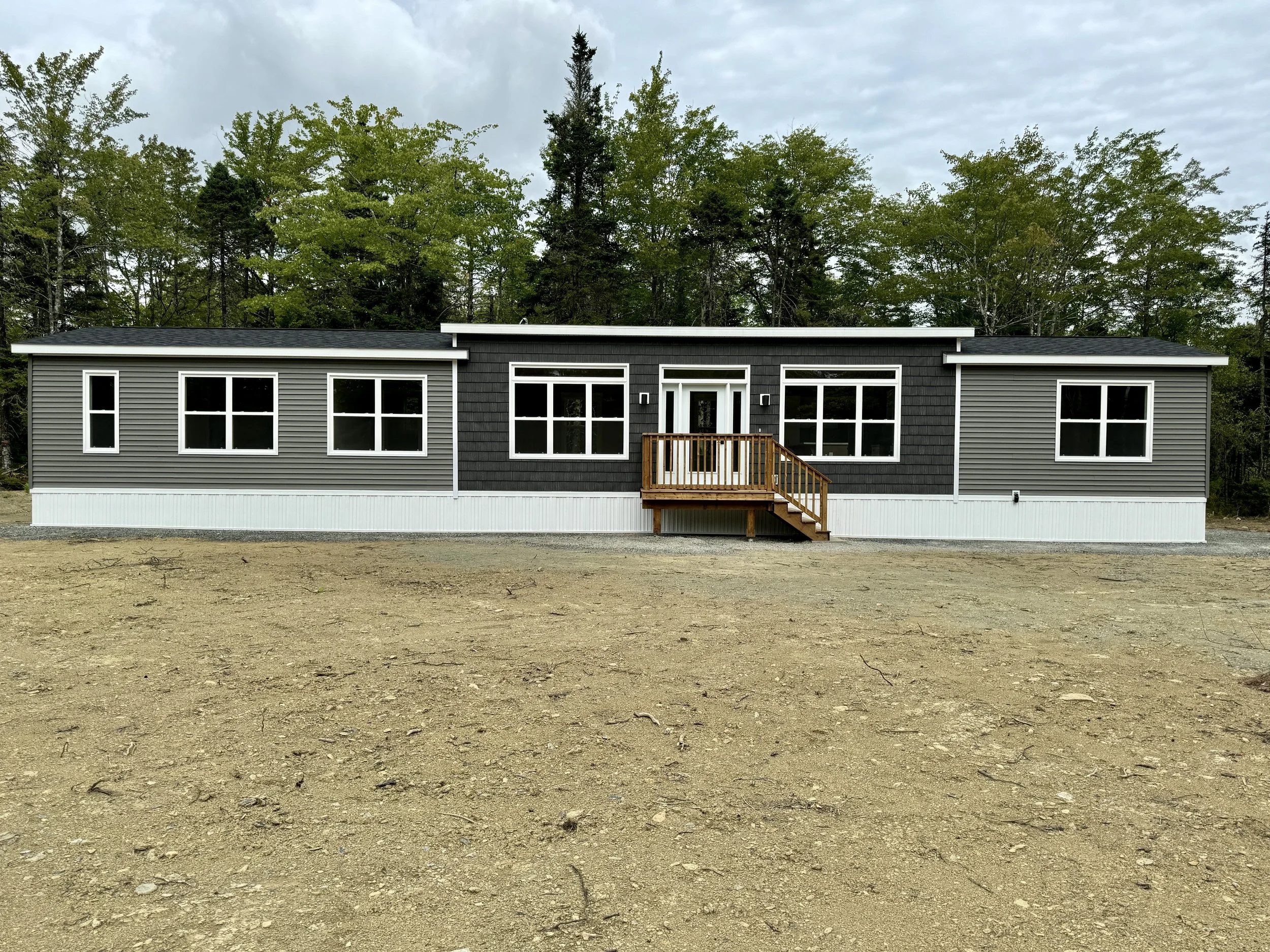 Newly built modern single-story house with gray and black siding, multiple windows, and a small wooden deck at the front, situated on a flat, cleared land with trees in the background.