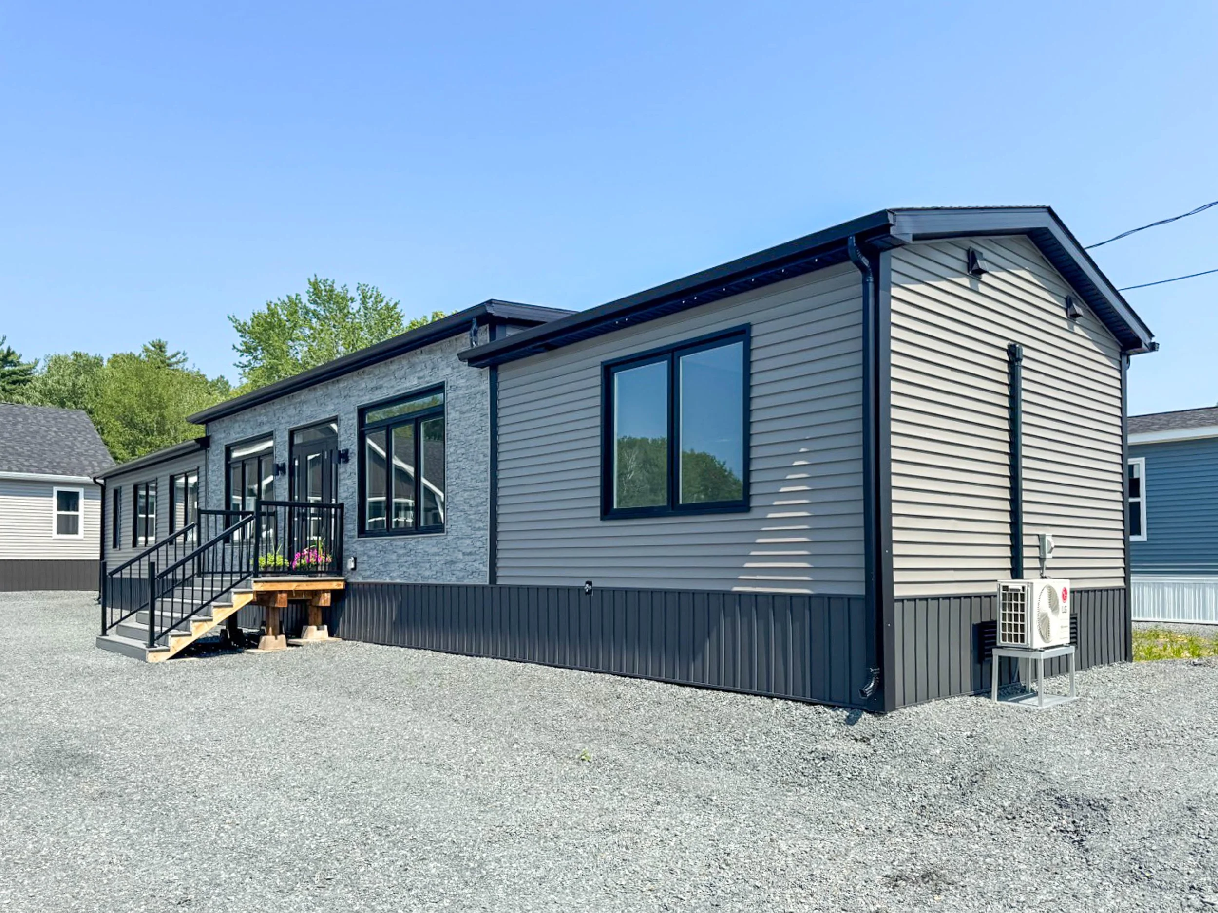Modern manufactured house with black framing, siding, and stone accents, situated on a gravel lot with a clear blue sky background.