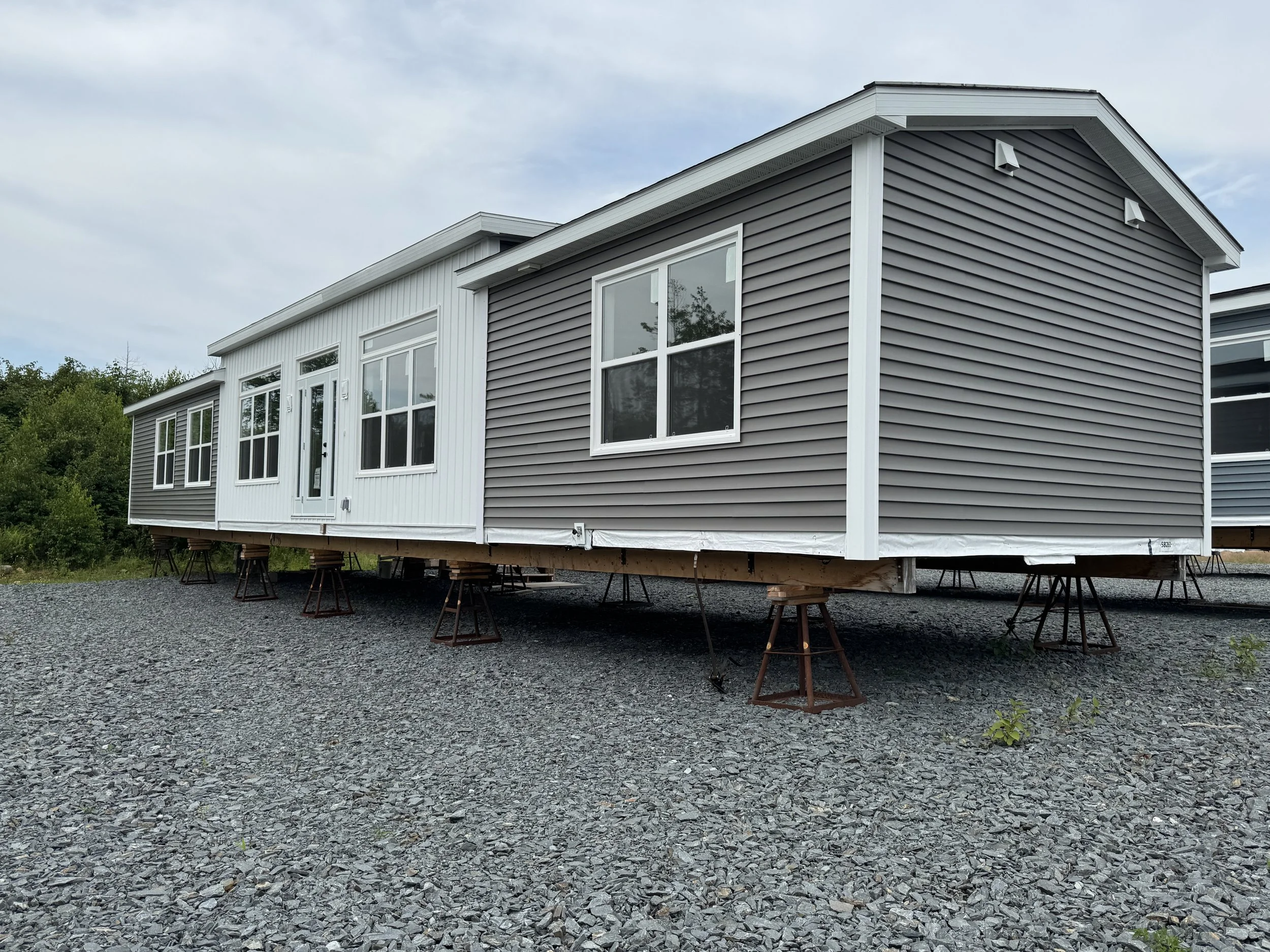 Exterior of a prefab house on elevated supports at a construction site with gravel and dirt ground, surrounded by trees and under a partly cloudy sky.