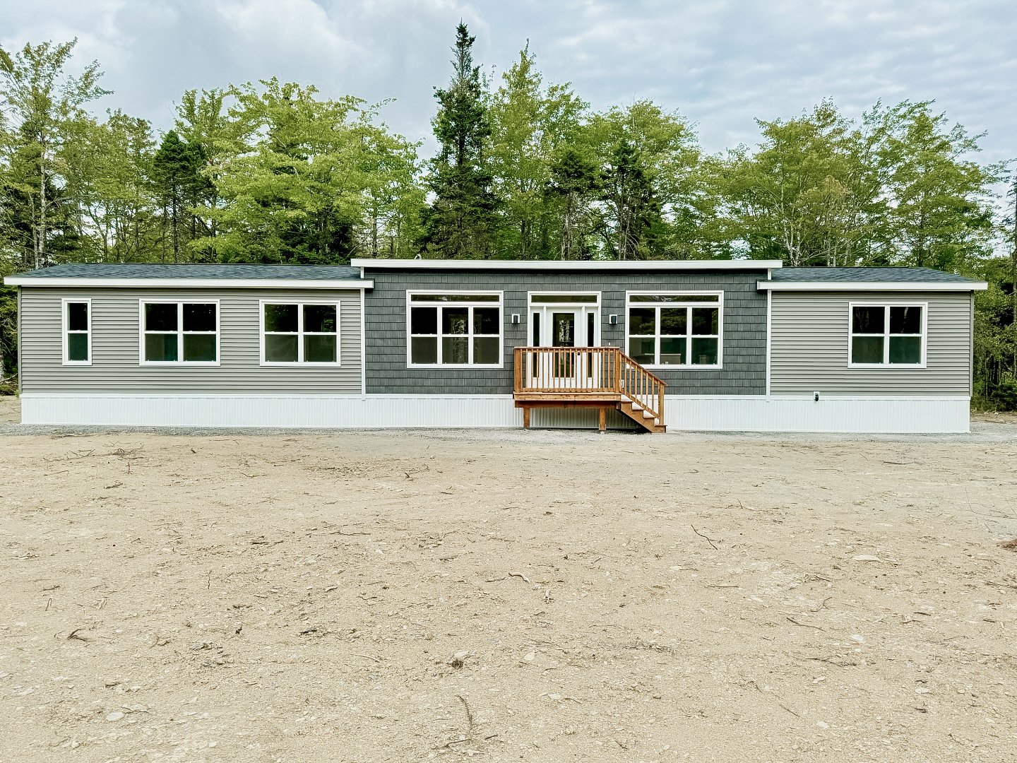 Newly built modern single-story house with gray and black siding, multiple windows, and a small wooden deck at the front, situated on a flat, cleared land with trees in the background.