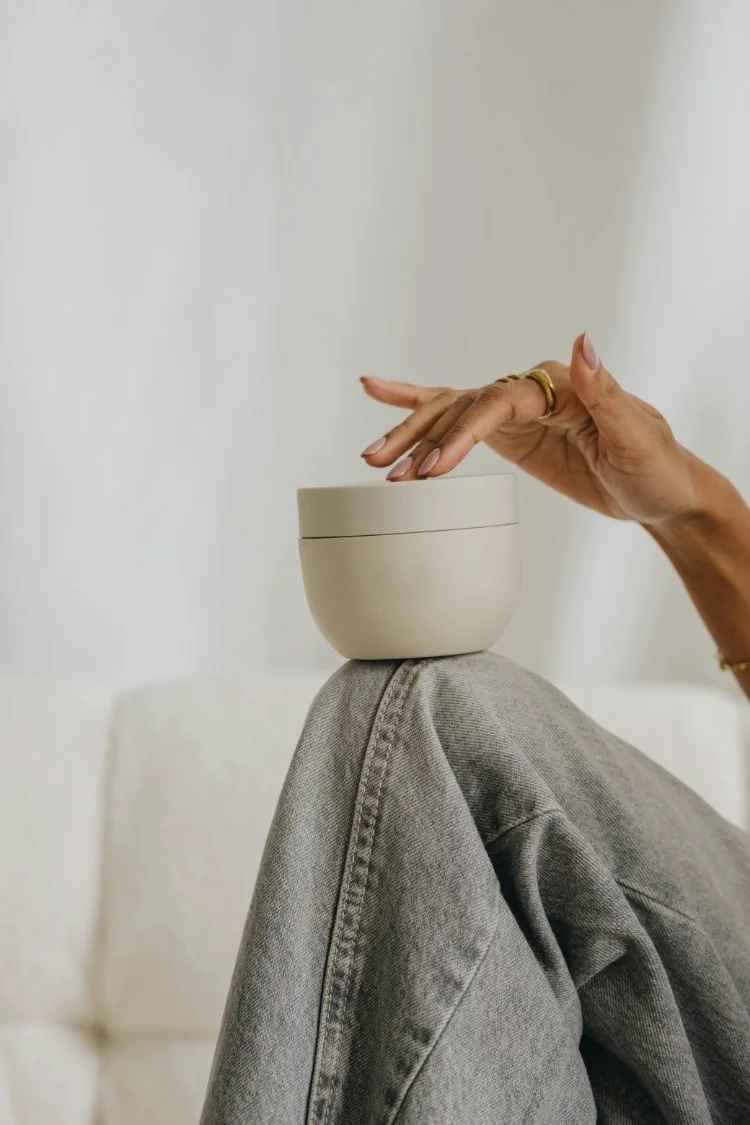 A person with painted nails and rings on their fingers balances a beige container on their knee, which is wearing light gray jeans, in a minimalist setting with a neutral background.