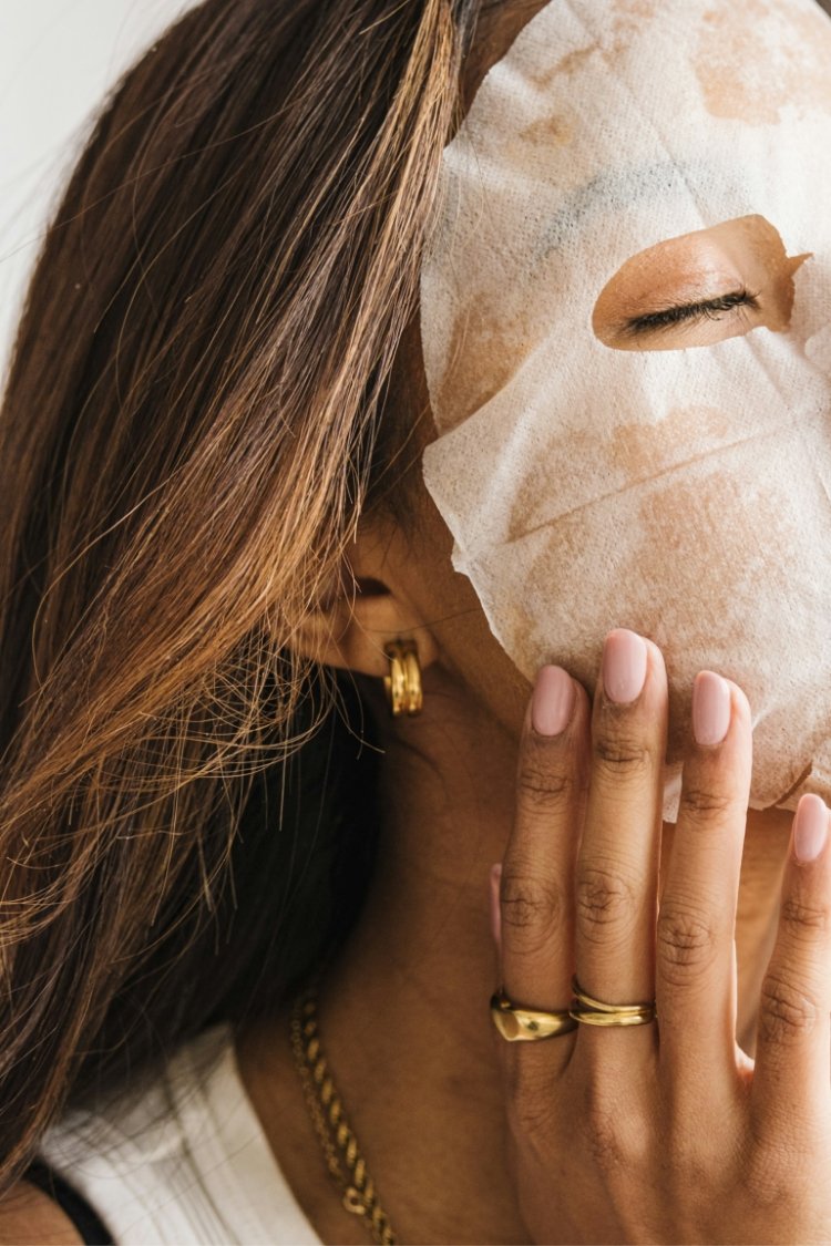 A woman wearing gold jewelry holds her hand near her face, which is covered by a facial mask and a protective sheet with eye and mouth holes, suggesting she is at a spa or skincare treatment.