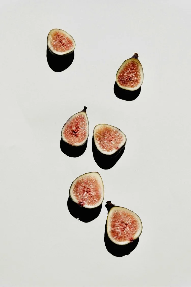 Six halved figs with pink interiors and greenish-yellow skin, arranged on a white background with shadows beneath each piece.