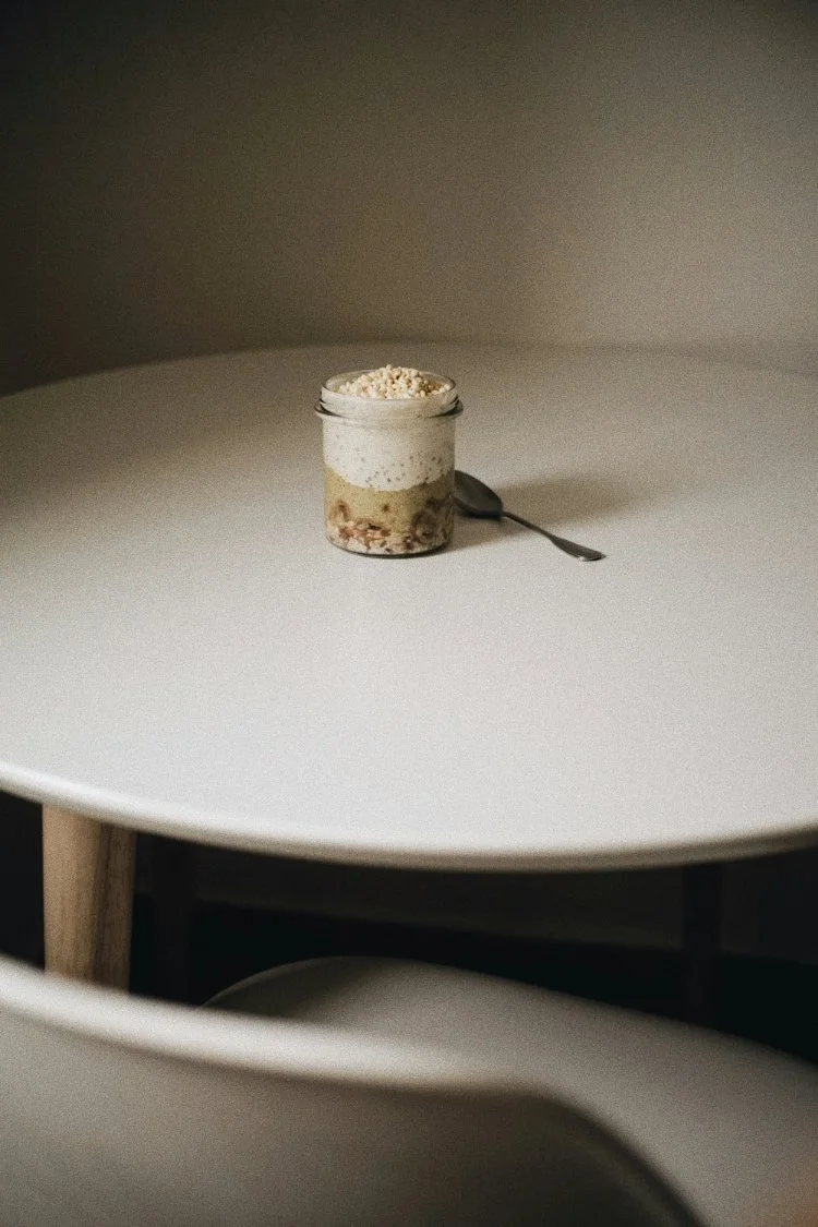 A small jar with layered dessert topped with whipped cream and white sprinkles, placed on a minimalist white table with a spoon beside it.