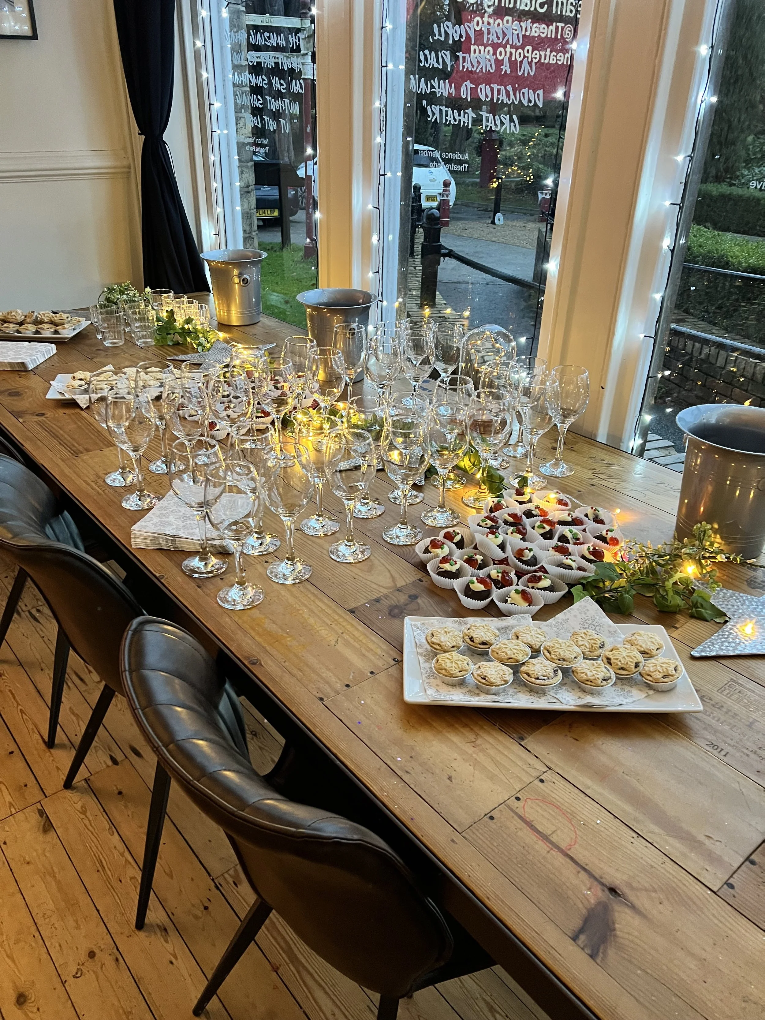 A wooden table set for a celebration with empty wine glasses, small desserts, and fairy lights near a window with a view of the outside.