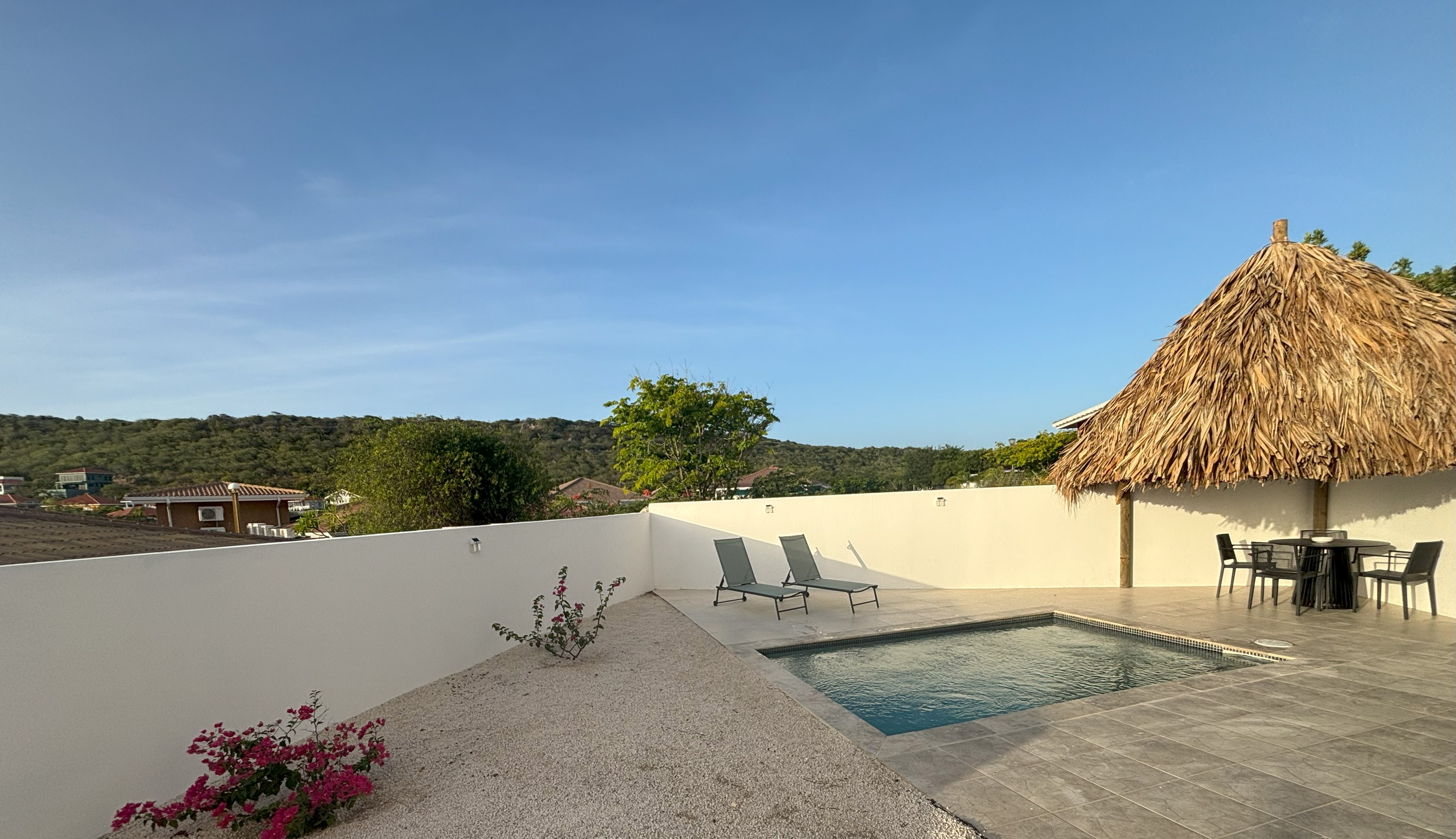 Porch with two black chairs, small black side table, and pink pillow, overlooking green hills and neighborhood houses under a partly cloudy sky.