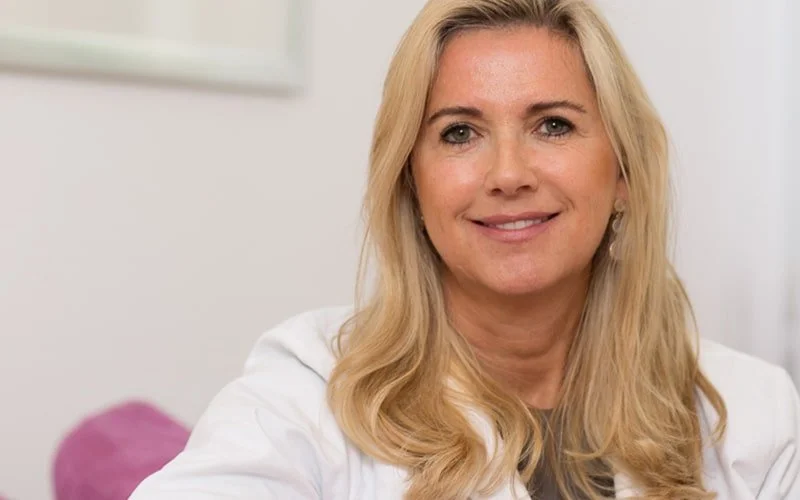 Porträt einer lächelnden Frau mit langen blonden Haaren in einem weißen Hemd, sitzend in einem hellen Raum.