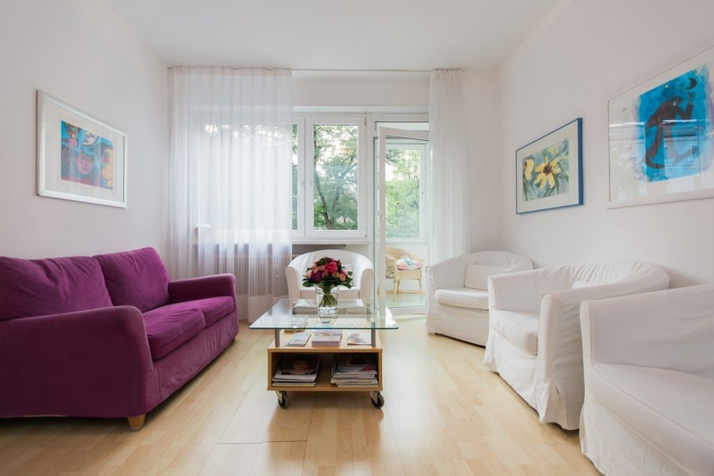 Helle Wohnzimmer mit weißen Wänden, mehreren Sesseln, einem großen Fenster, einem Glastisch mit Blumen, und bunten Kunstwerken an den Wänden.