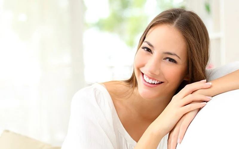 Lächelnde junge Frau sitzt auf einem Sofa, an einem hell erleuchteten Raum mit Fenstern im Hintergrund.