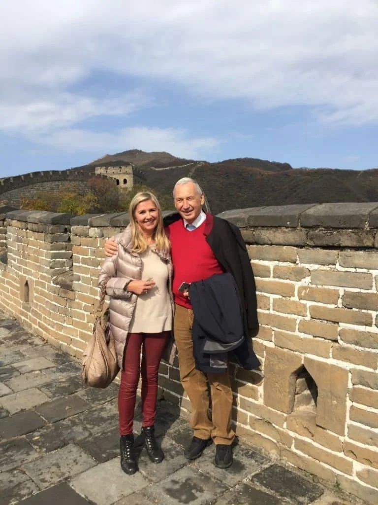 Ein Mann und eine Frau stehen lachend an einer alten Steinmauer mit dem Chinesischen Abschnitt der Chinesischen Mauer im Hintergrund, bei schönem Wetter und blauen Himmel.