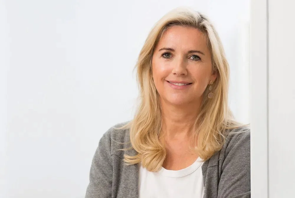 Frau mit blonden Haaren, grauem Blazer und weißem Oberteil posiert vor weißem Hintergrund.