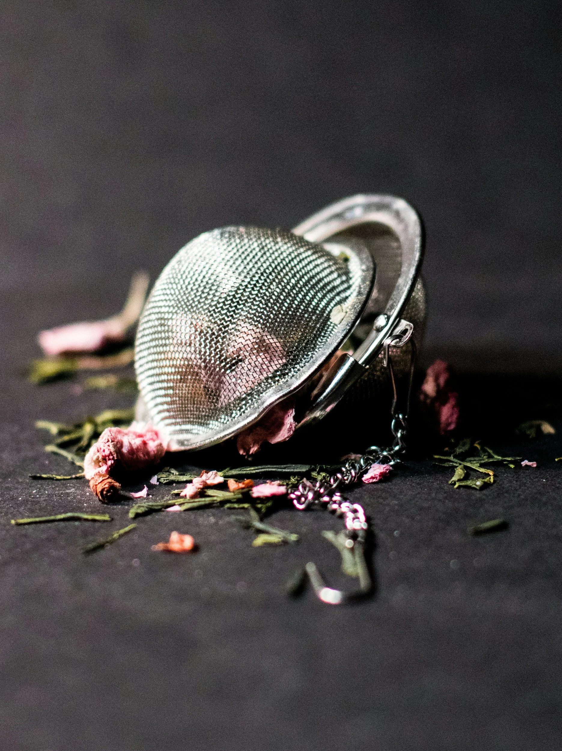 A metal tea infuser surrounded by dried pink flower petals and green leaves on a dark surface.