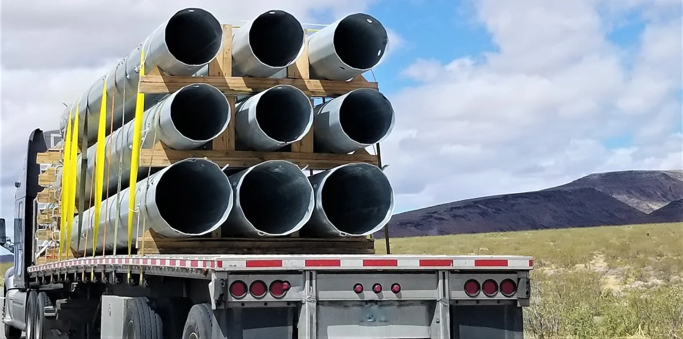 Flatbed truck transporting large metal pipes in an open landscape with hills and cloudy sky.
