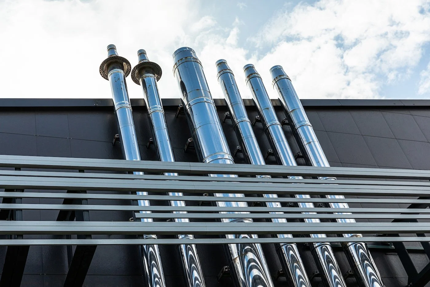 Multiple large, shiny metal pipes mounted on a black building exterior under a cloudy sky.