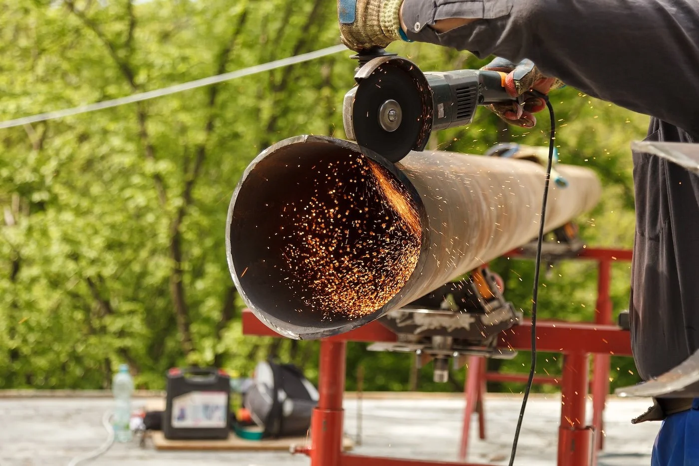 Person using an angle grinder to cut through a metal pipe, with sparks flying, outdoors with green trees in the background.