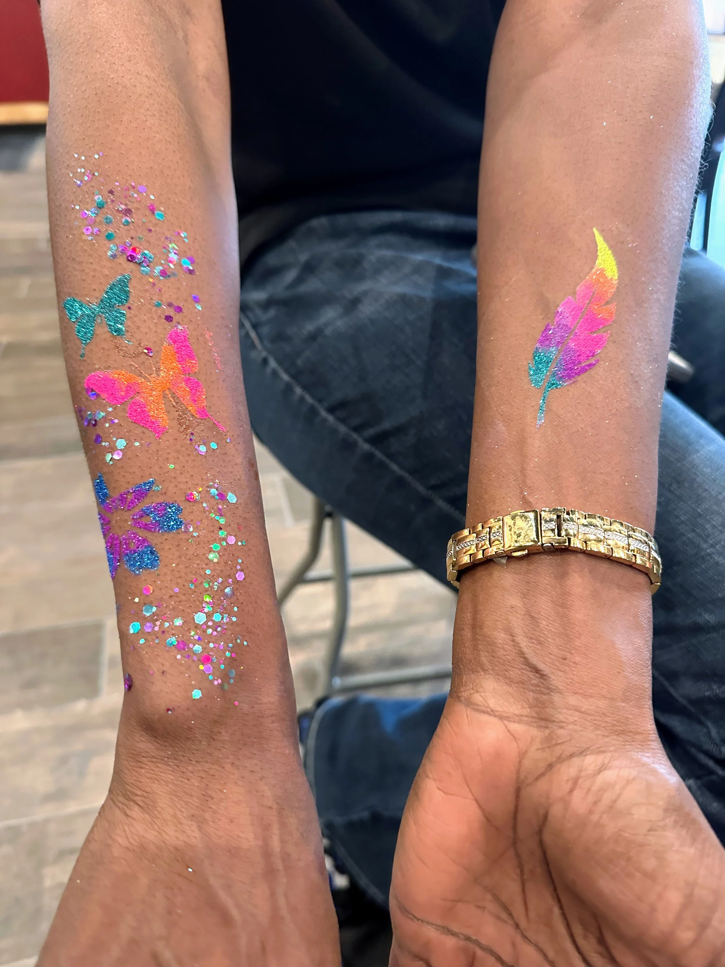 A person of color shows their arms with brightly colored feather and butterfly glitter tattoos