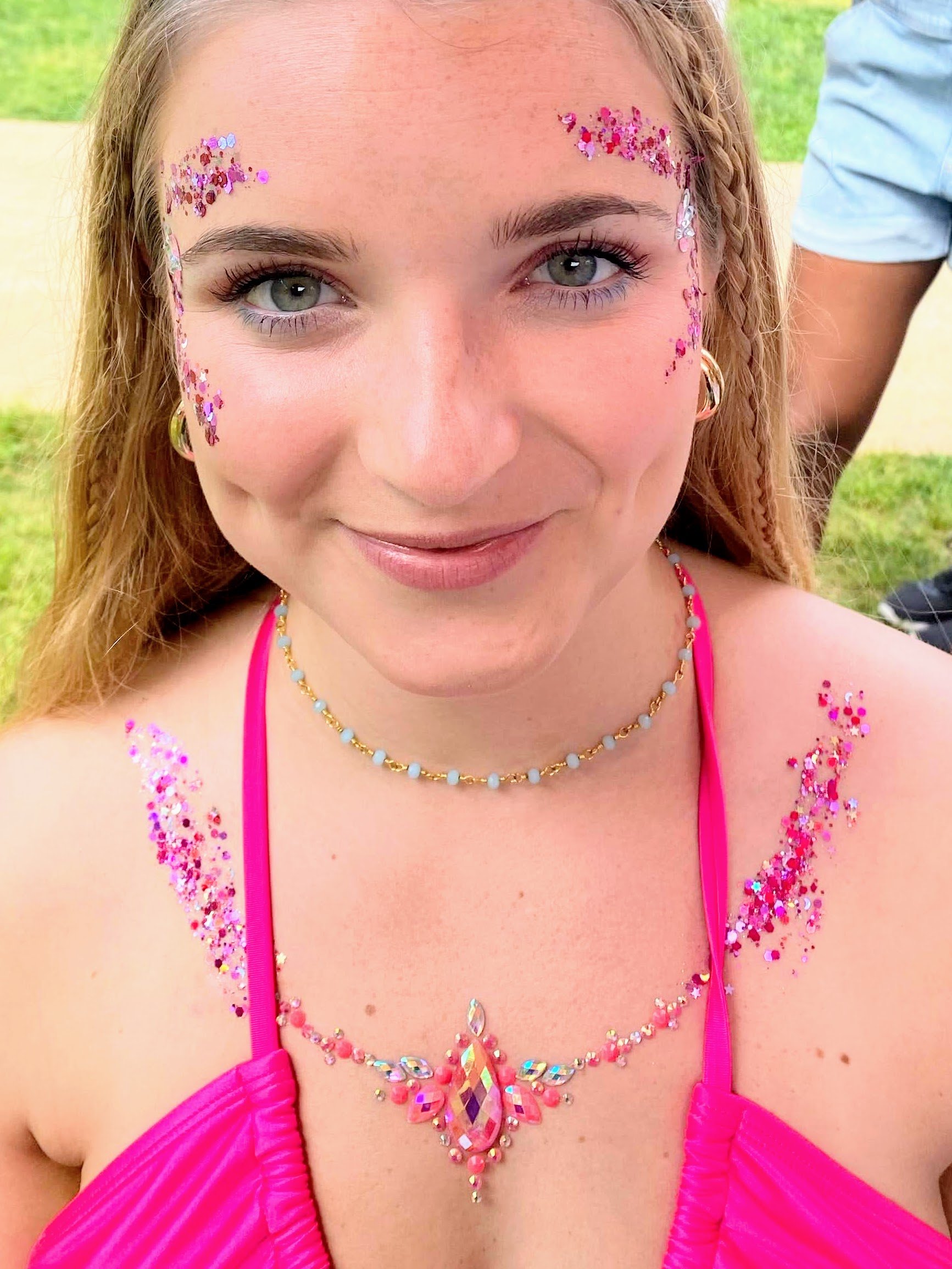 A college age girl smiles with her face and chest adorned with bright pink glitter and fancy gems