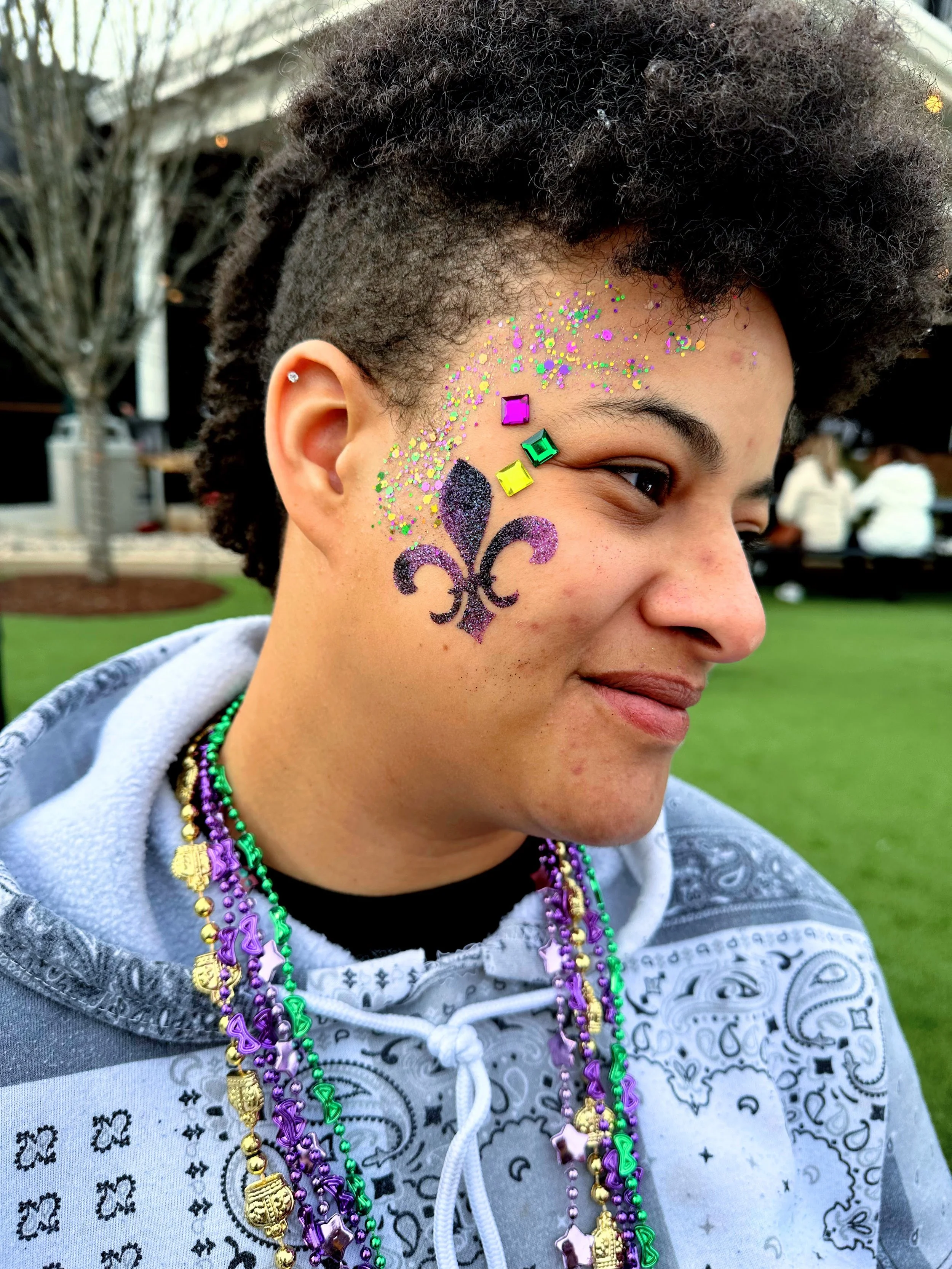 A young woman of color smiles and tilts her head to show off her elaborate glitter pattern and fleur de lis glitter tattoo on her cheek