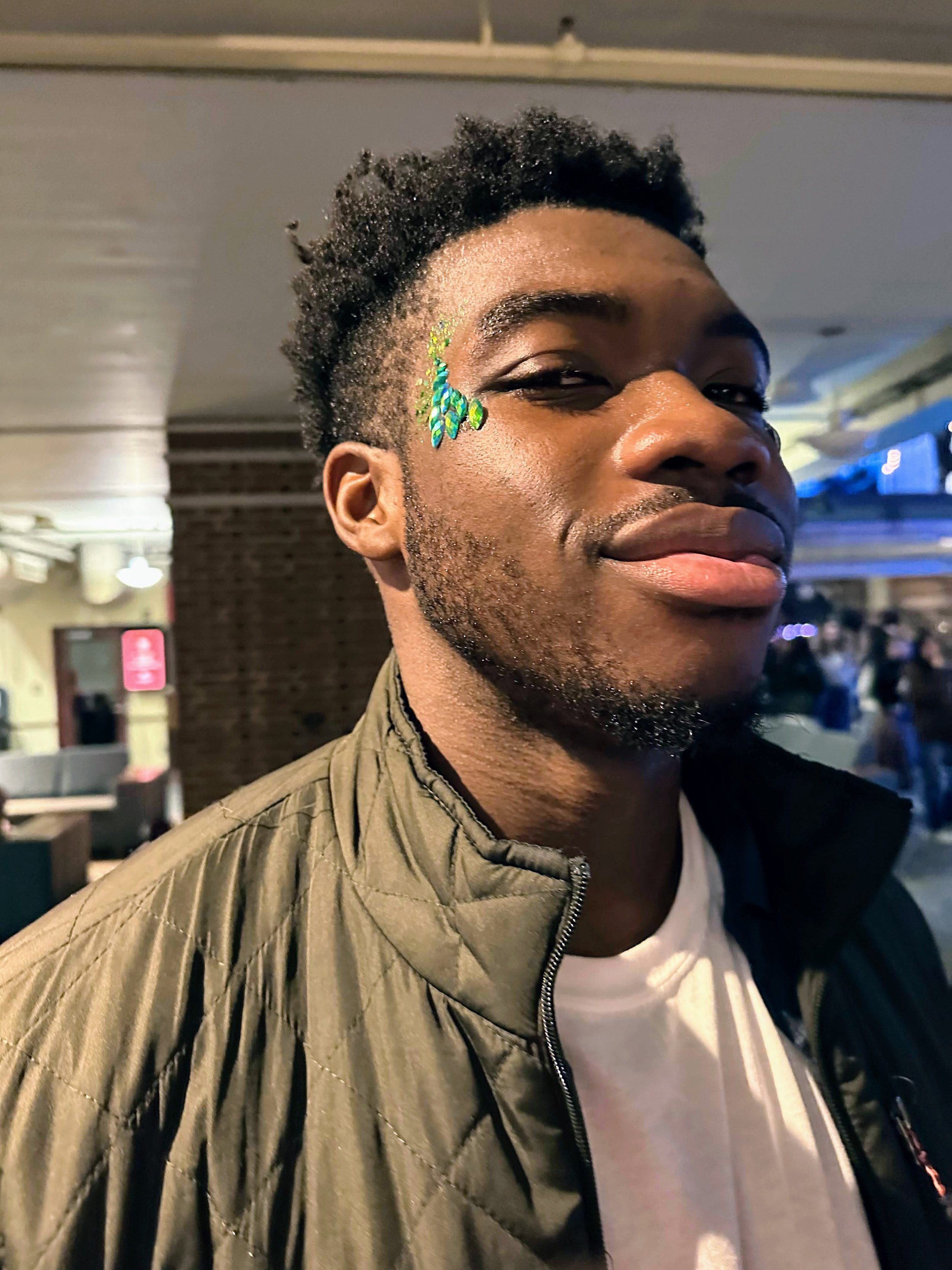 A young man of color smirks into the camera with bright green glitter designs adorning the side of his face