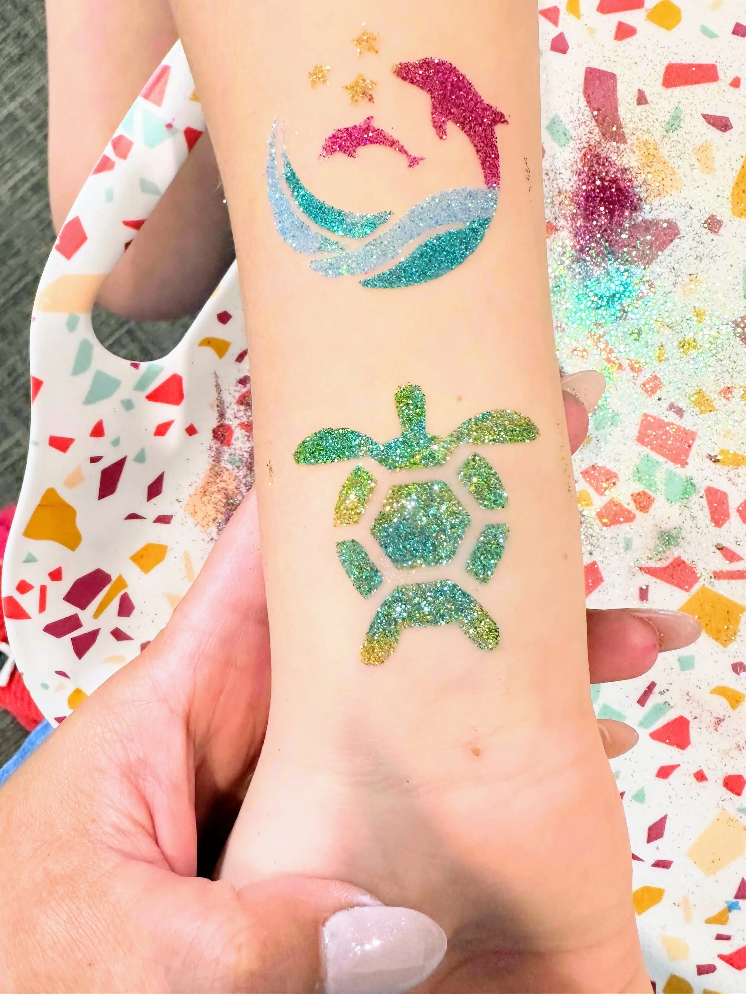 A young person extends their arm to show off brightly colored dolphin and sea turtle glitter tattoos