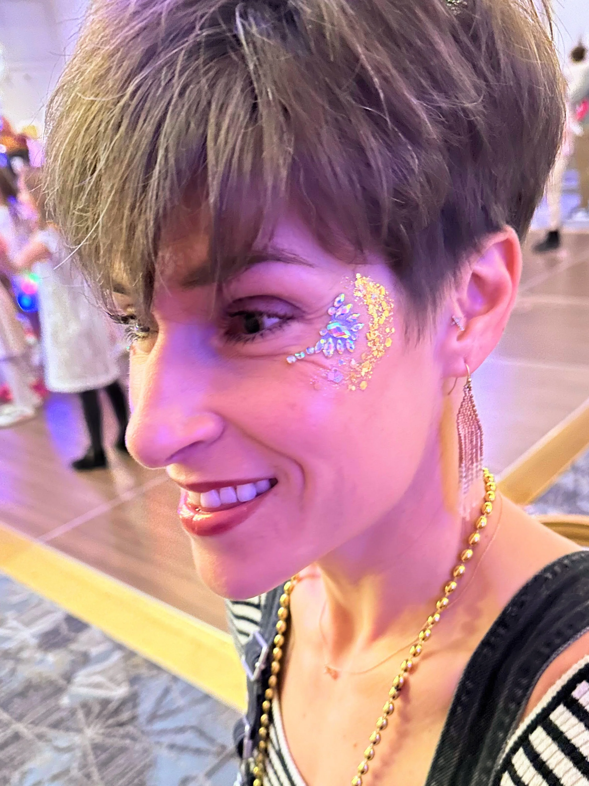 A woman with short blond hair smiles with her face decorated with blue and yellow glitter patterns