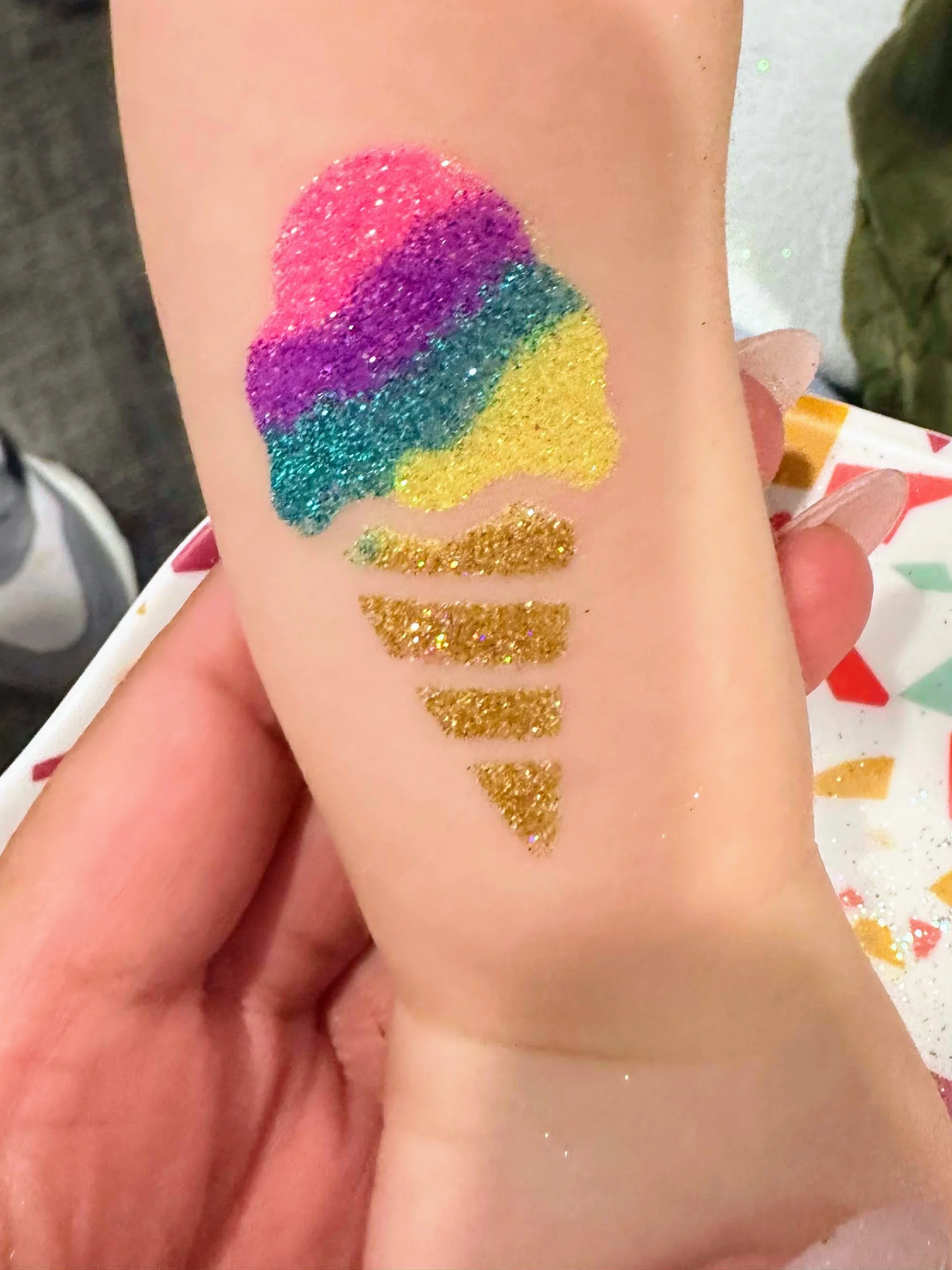 A young child's arm shows off a rainbow colored ice cream cone glitter tattoo