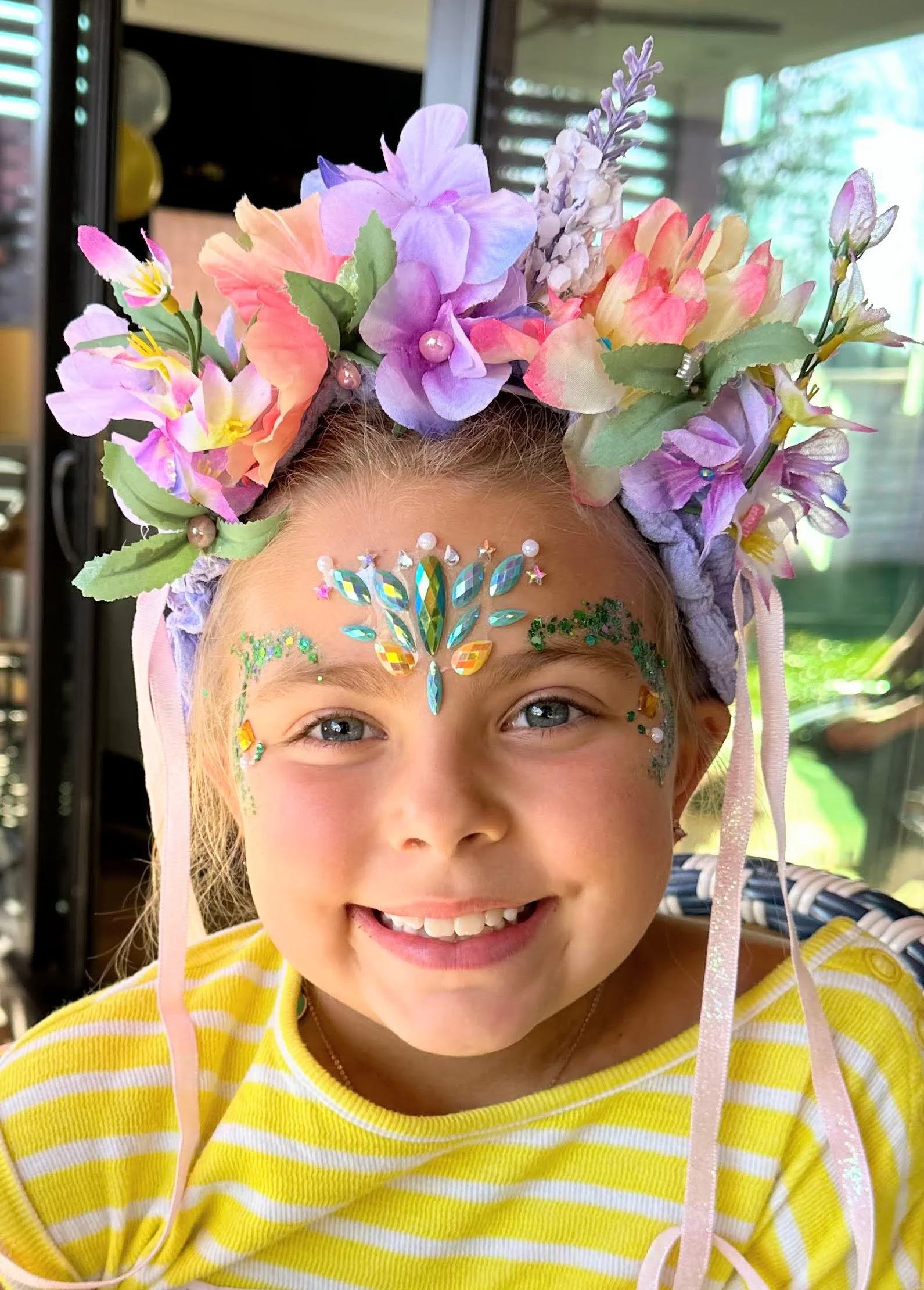 A young girl with a flowered crown smiles with her face adorned with bright glitter and gems