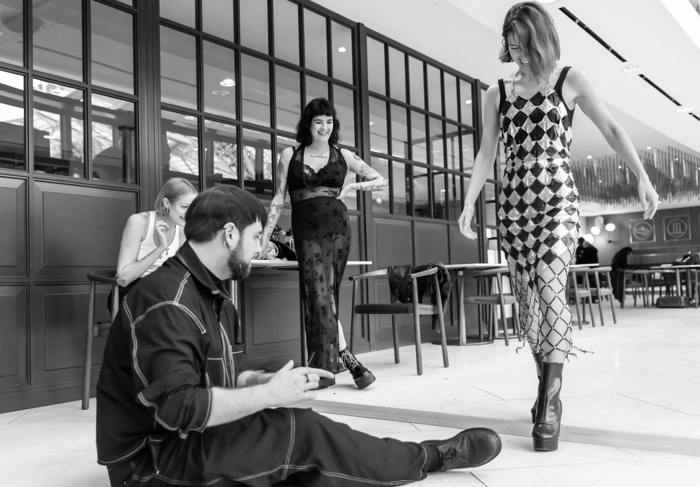 A woman in a patterned dress and platform boots models as two women and a man observe and smile in a modern interior space.