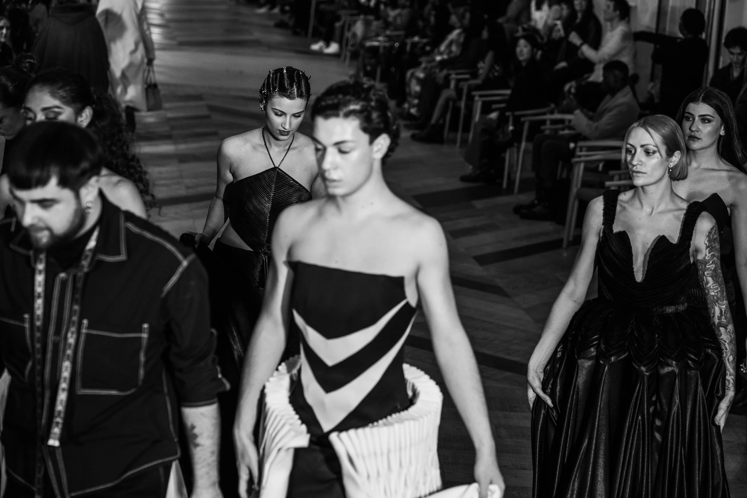 Models walking in a fashion show, wearing black and white designer dresses, with an audience seated in the background.