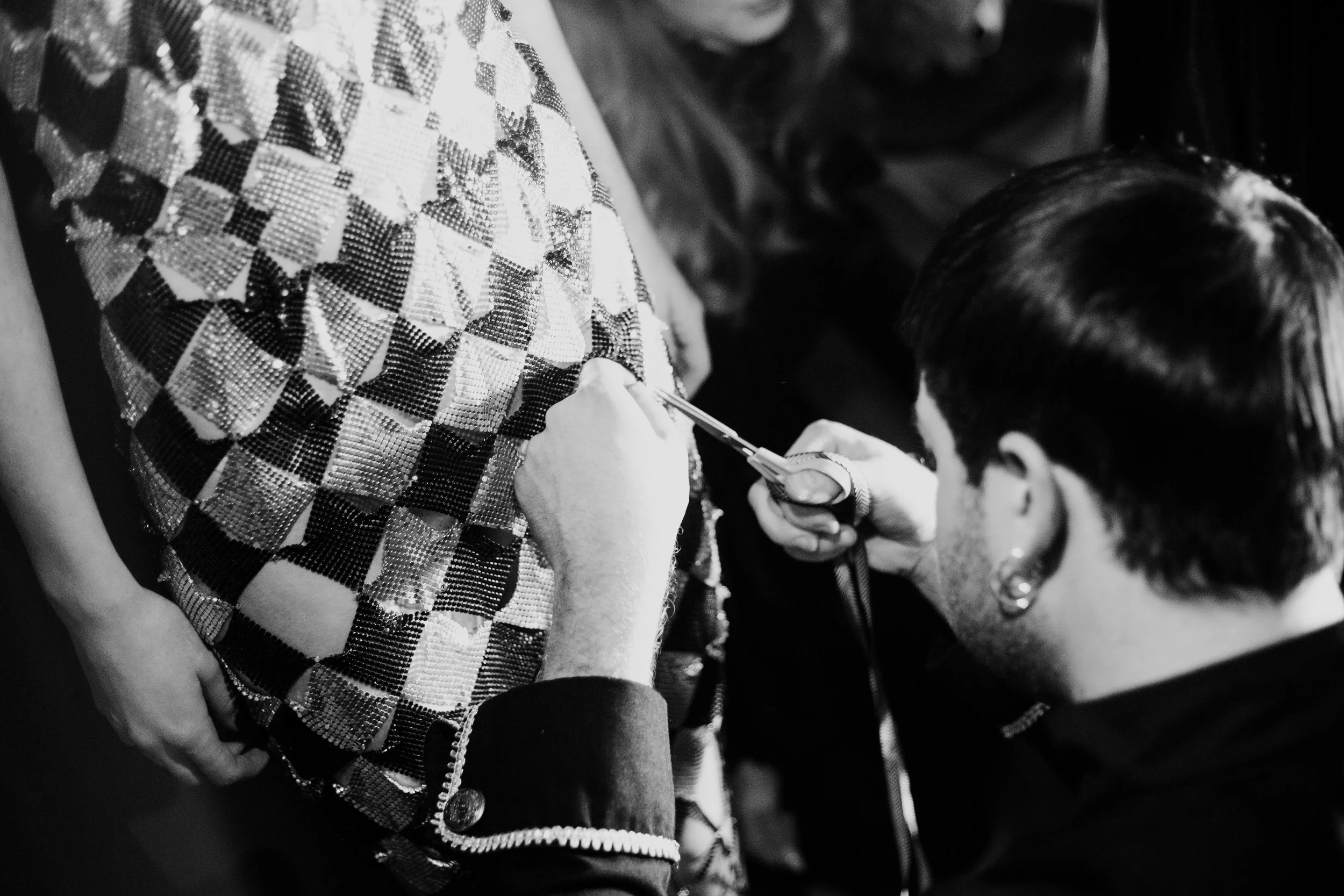 A person is sewing or tailoring a checkered fabric garment while another person watches closely.