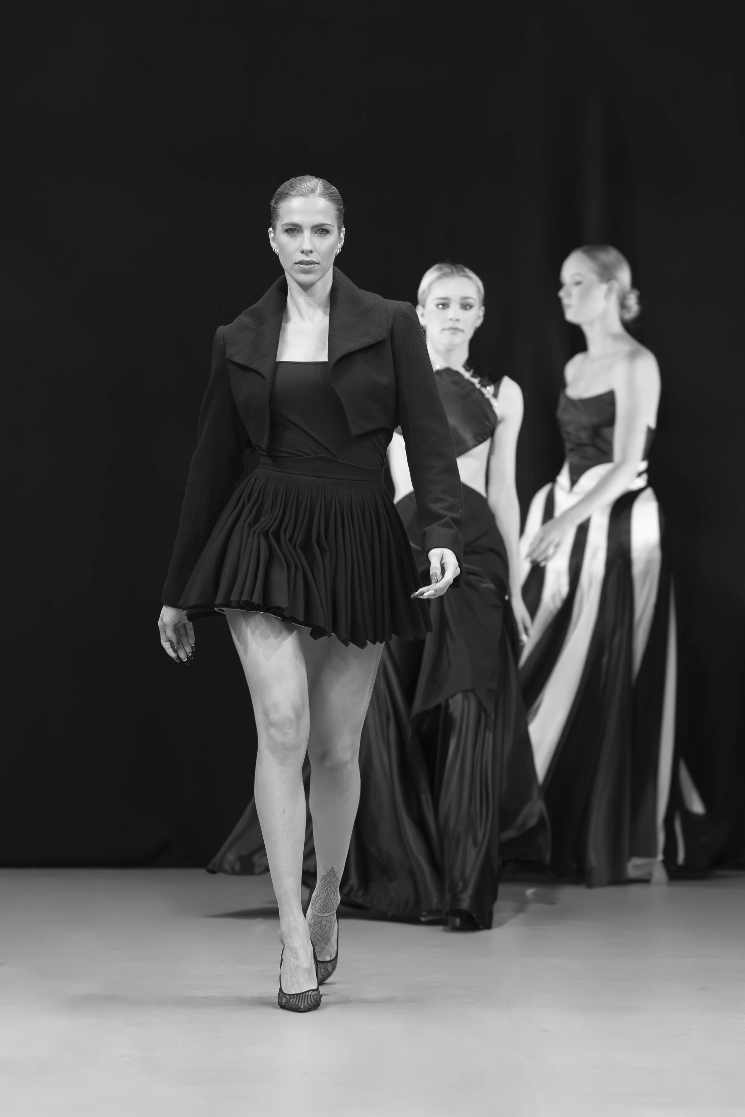 Black and white photo of three female models on a runway, dressed in stylish evening wear. The front model wears a dark jacket over a pleated skirt, with high heels and an ankle tattoo. The two models in the background wear elegant, long, black and white gowns.