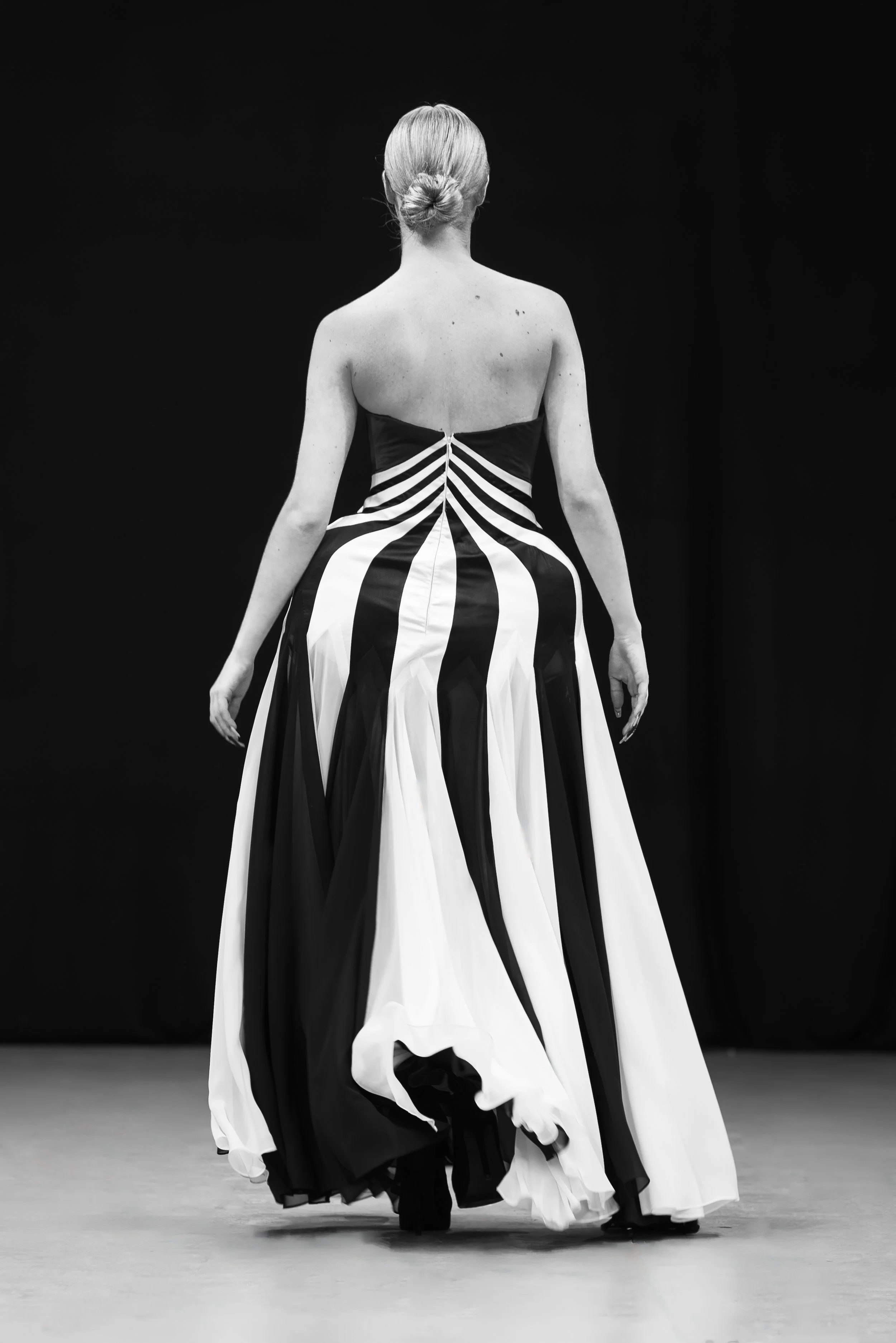 Woman walking on runway with long, striped black and white dress, hair in a bun, back view, black background.