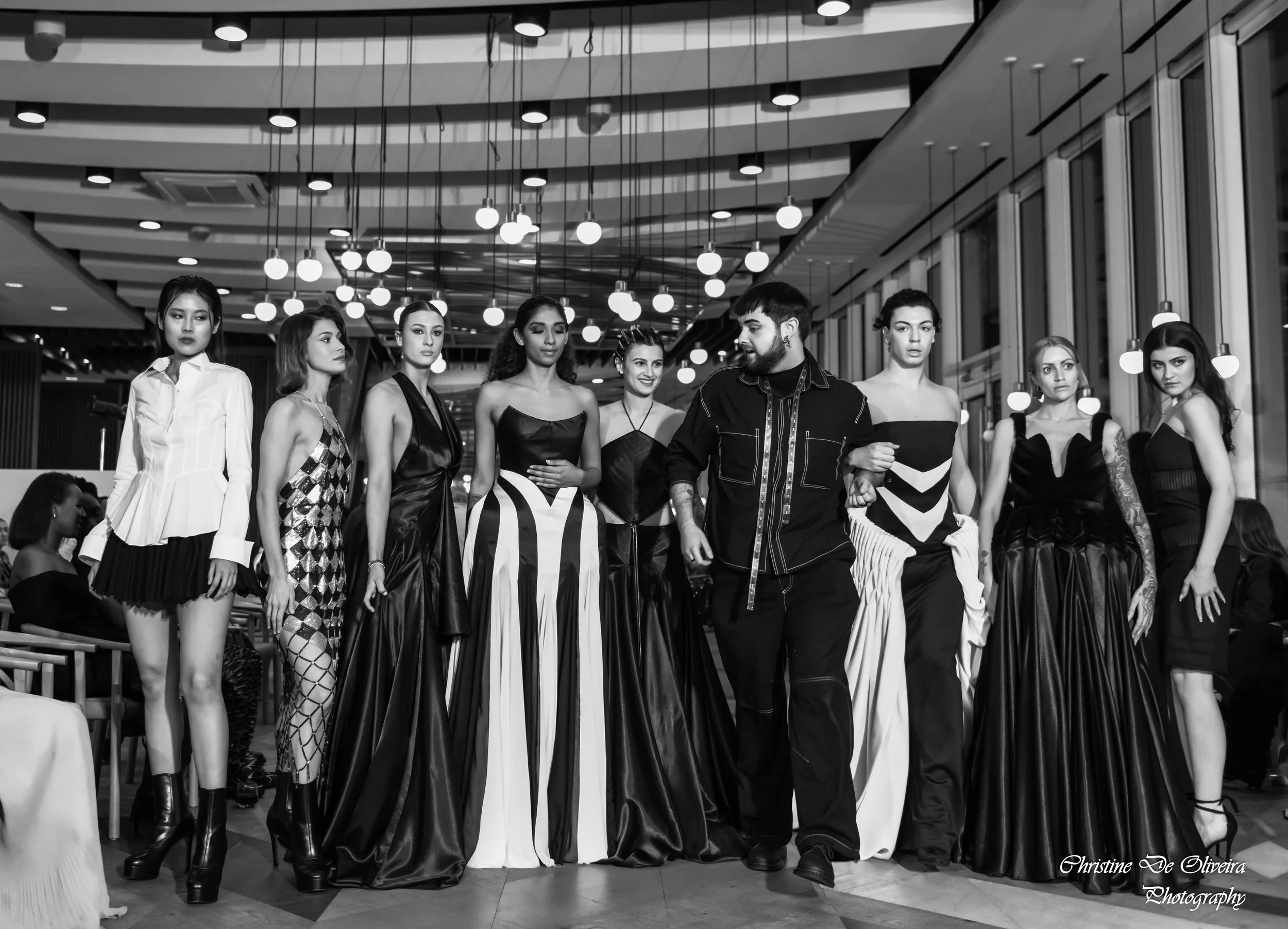 Black and white photo of a fashion show with nine models in evening gowns standing in a line while a man walks beside them, inside a modern venue with hanging lights and large windows.