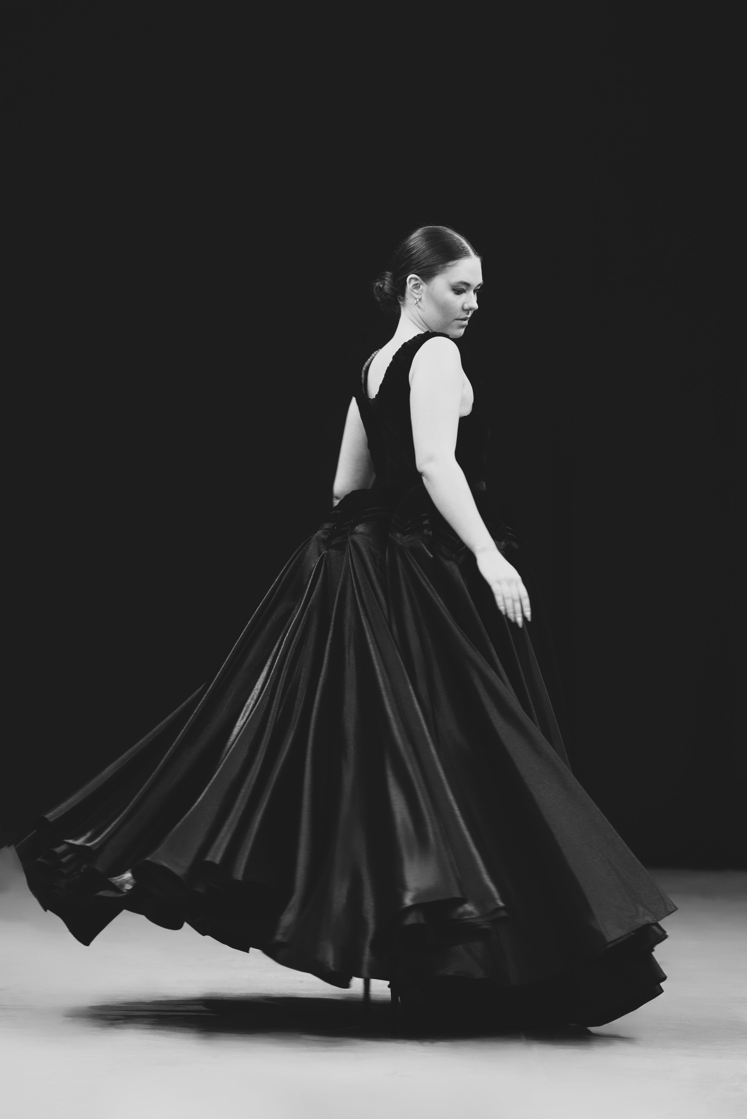 A woman in a long, voluminous black gown walking on a stage, captured in black and white.