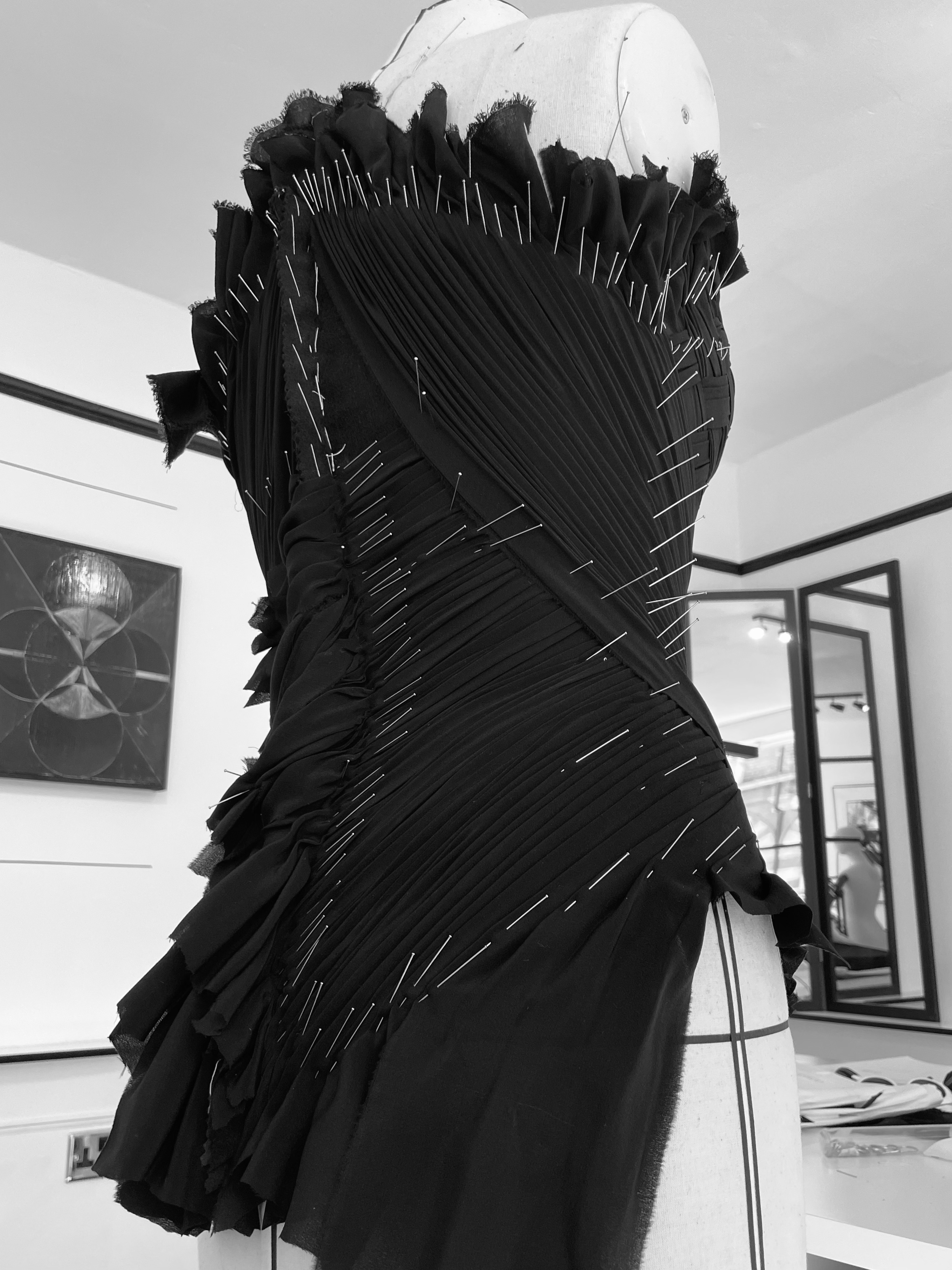Black dress draped on a mannequin, with numerous sewing pins inserted into the fabric, positioned in a sewing or design studio.