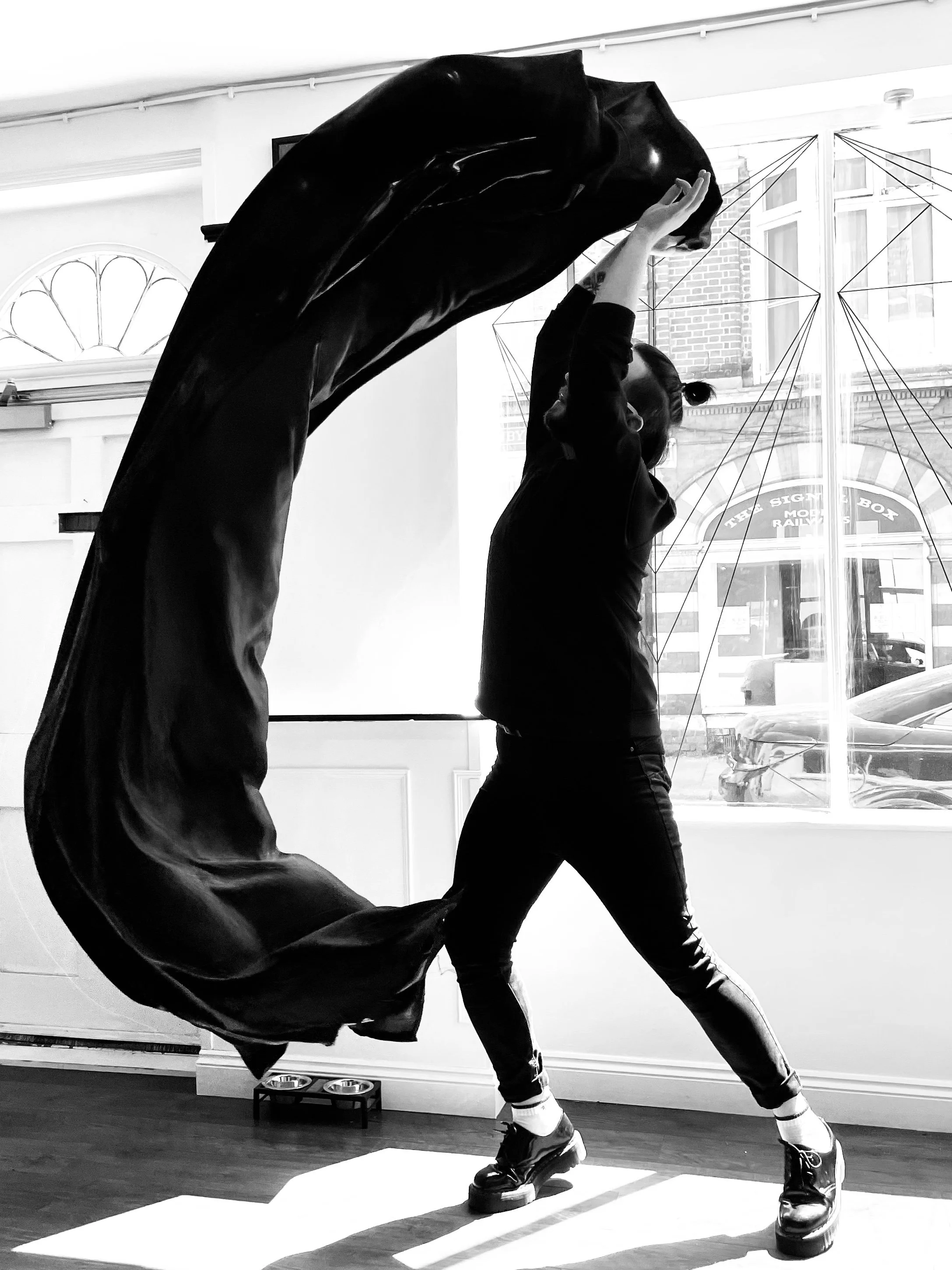 Person with hairstyle in buns is dancing, lifting their large piece of fabric in a sweeping motion, in front of a large window with city view, in black and white.