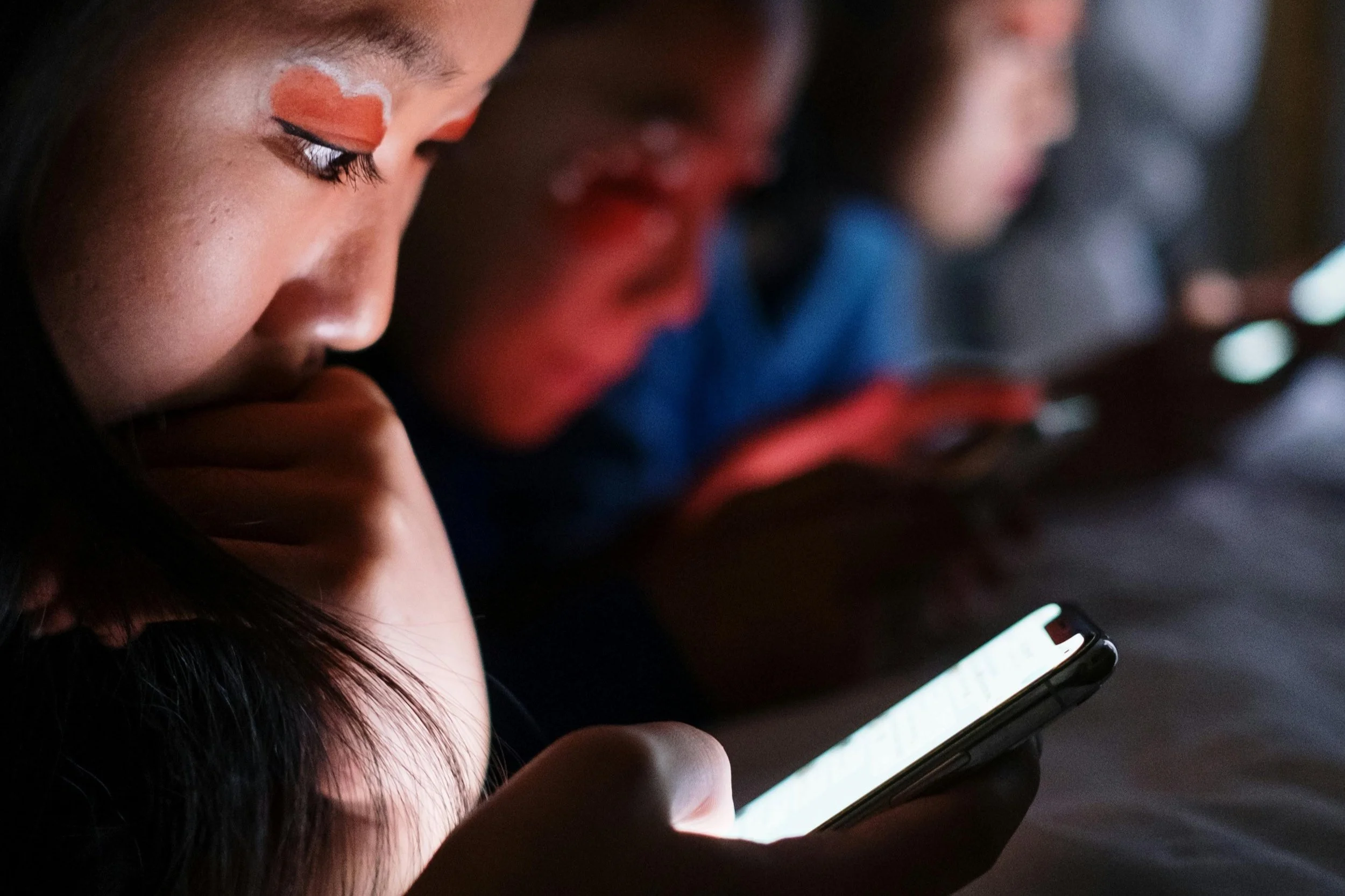 Nahaufnahme von einer Person, die auf ein Smartphone schaut, im Hintergrund weitere Menschen in einem dunklen Raum.