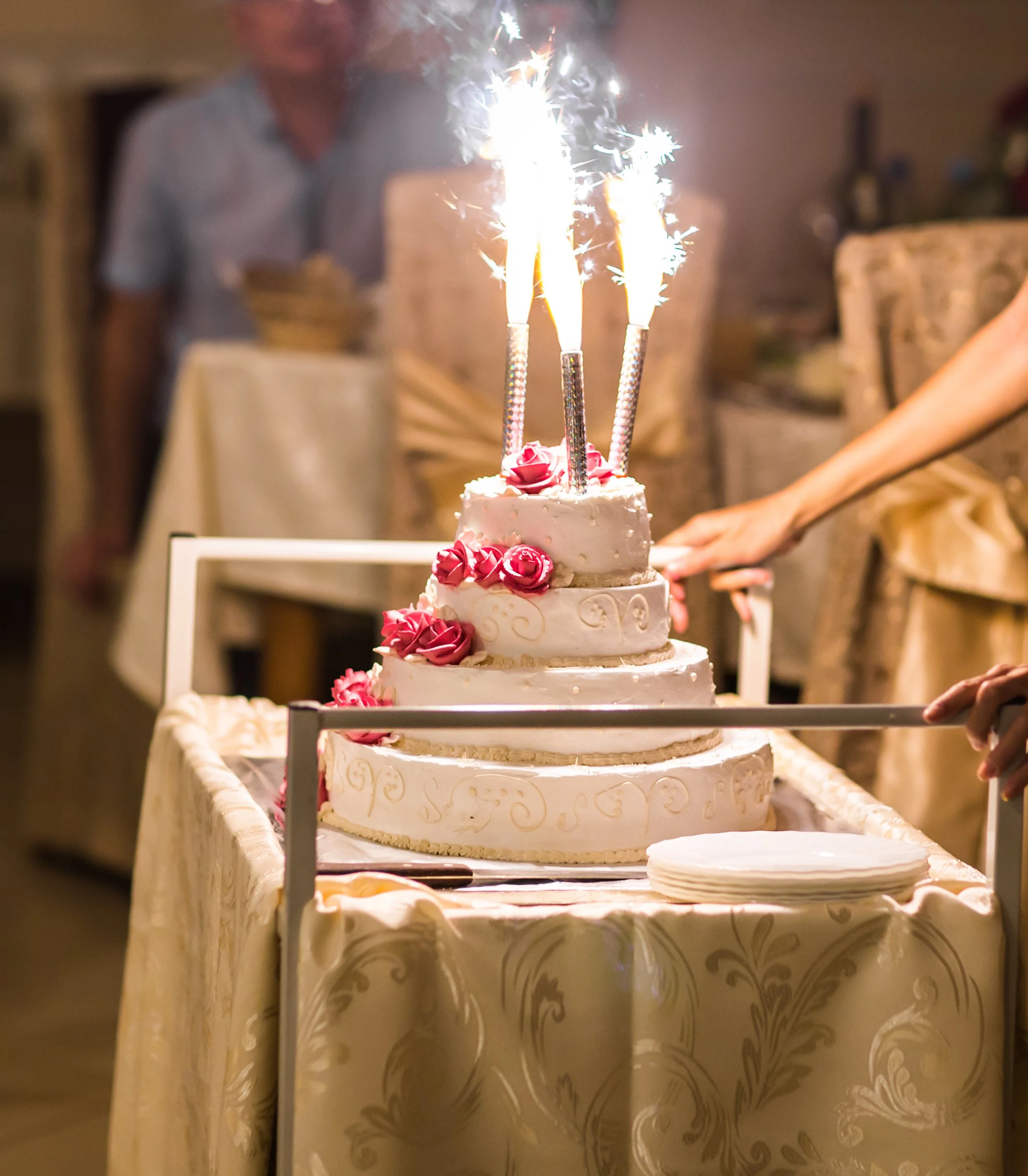 A multi-tiered wedding cake with pink roses and sparklers on top, placed on a decorated table at a celebration.