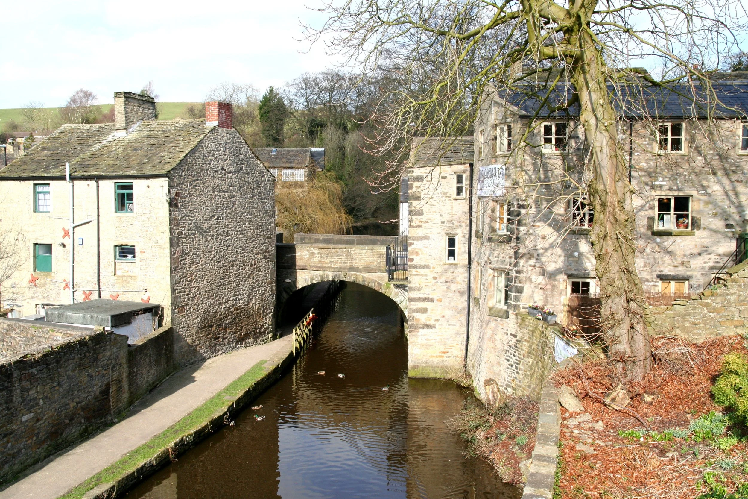 A historic stone bridge over a small river, with old stone buildings on either side and a leafless tree in the foreground.