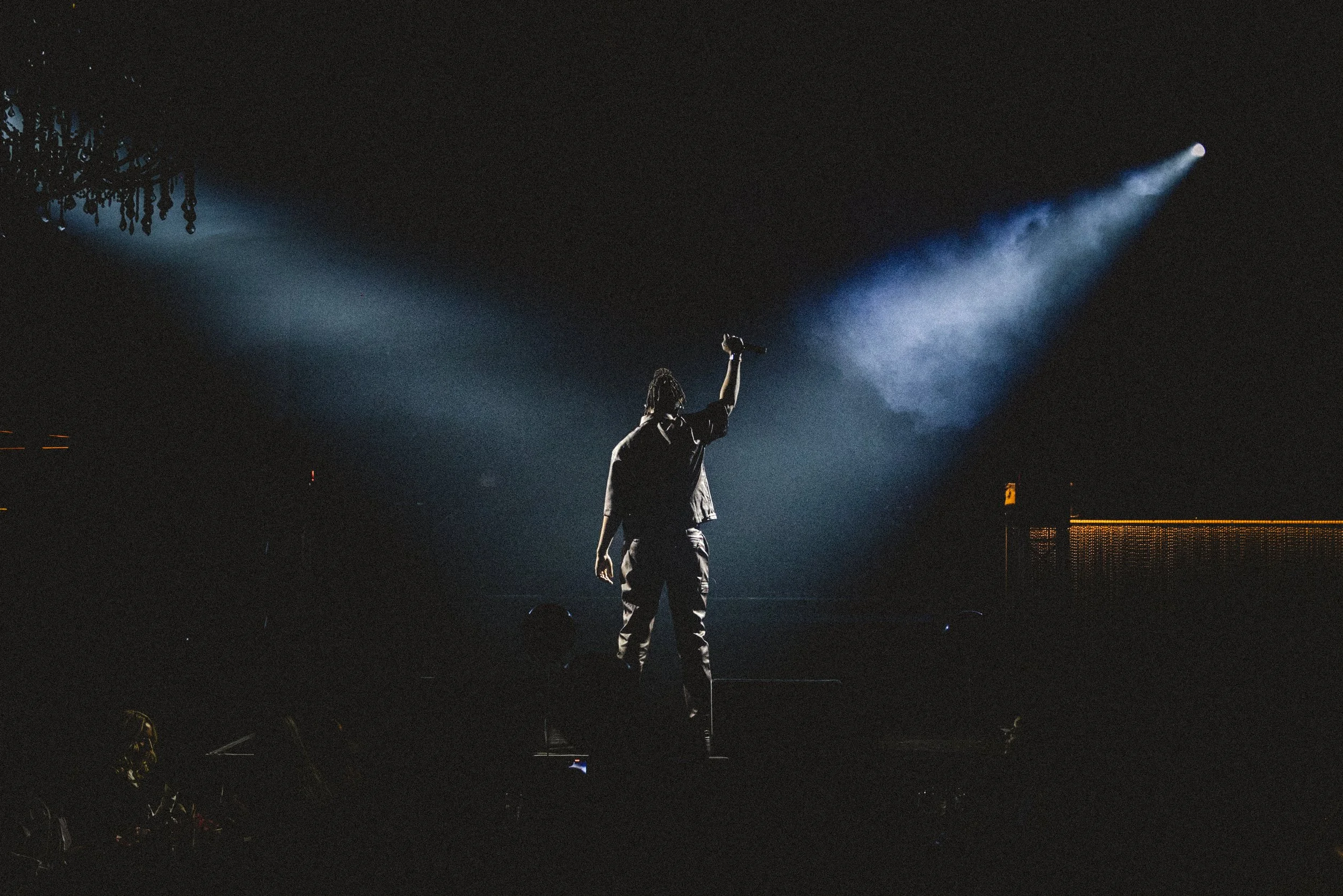 Performer on stage holding microphone, illuminated by spotlight in dark venue with chandelier overhead.