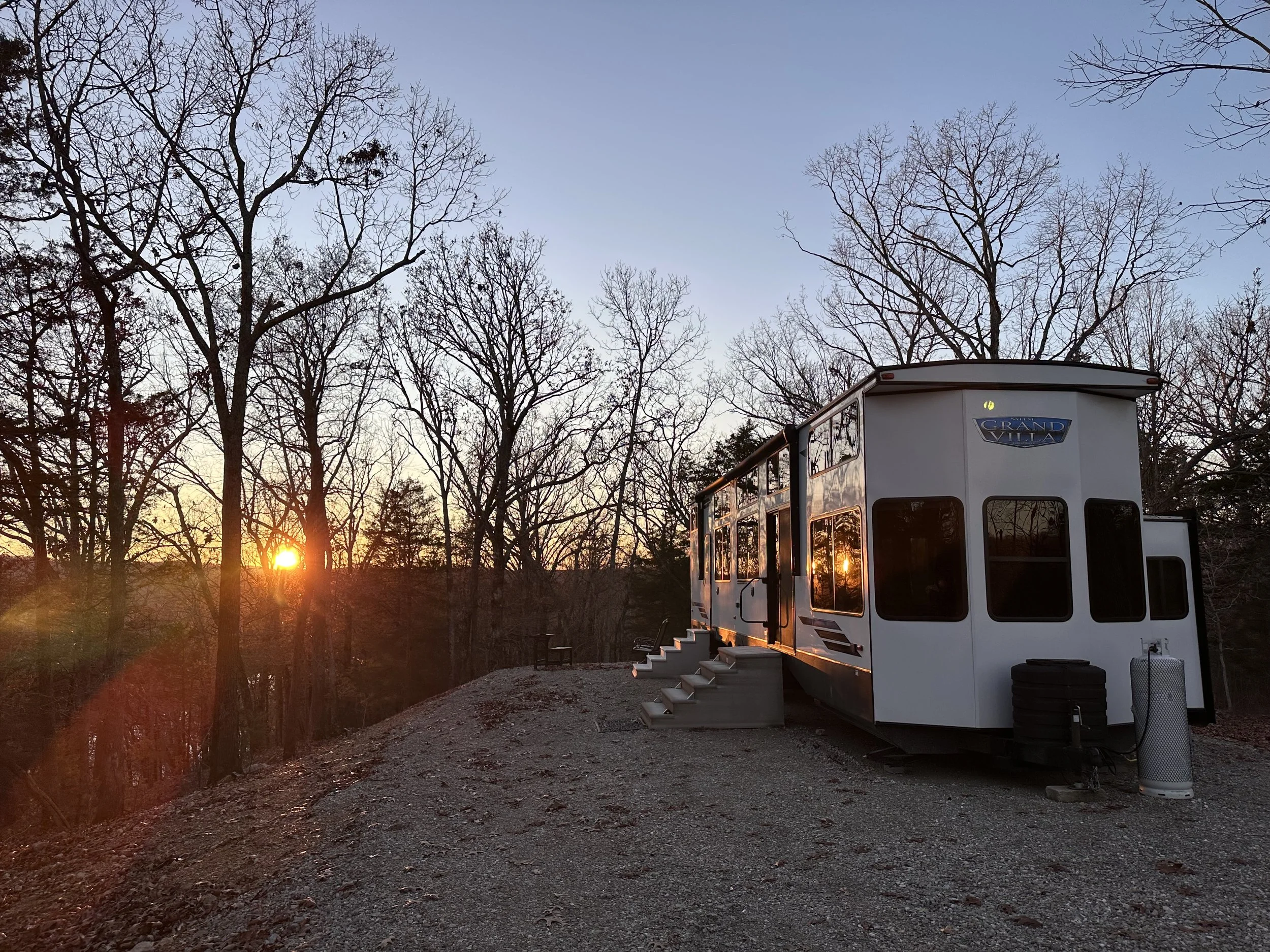A large white recreational vehicle (RV) with tinted windows parked outside a metal building during sunset, with propane tanks in front.