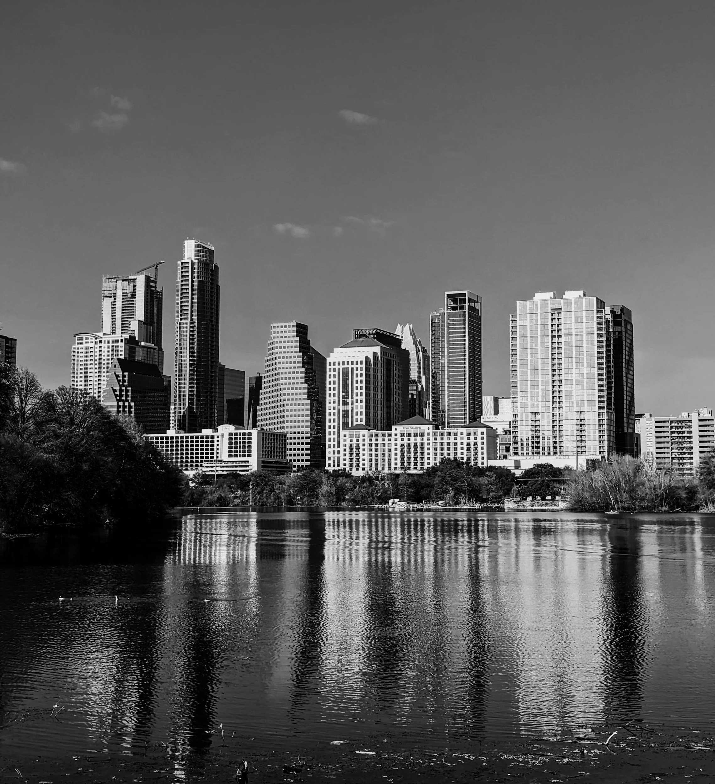 Black and white photo of a Austin skyline with tall buildings reflecting on a body of water in the foreground, trees along the shore, and scattered clouds in the sky.