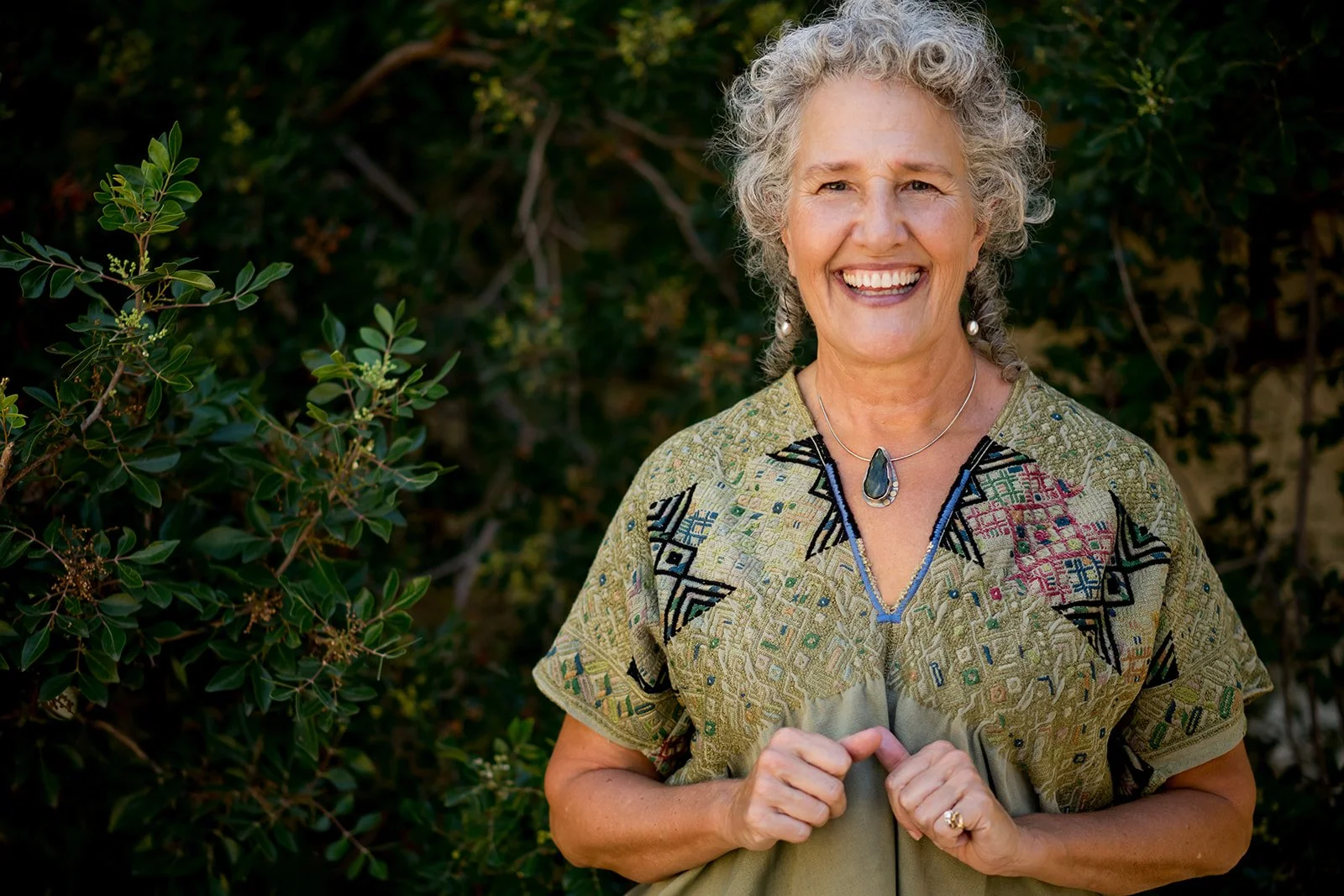Best Executive coach, Ellen Lindsey with curly gray hair smiling outdoors, wearing a patterned top with a large pendant necklace, standing next to green bushes.