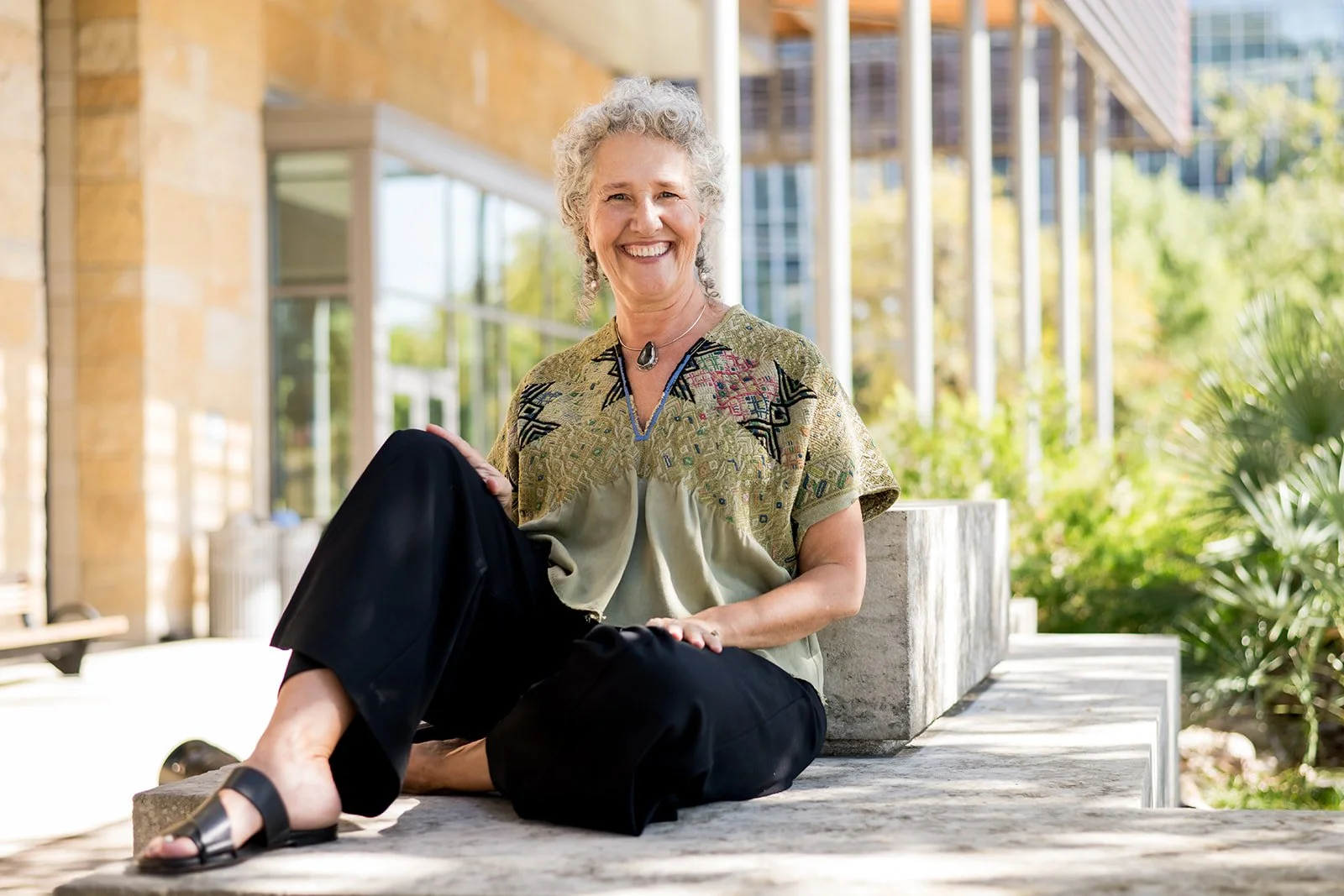 Ellen Lindsey, executive coach in Austin, with curly gray hair, smiling and sitting cross-legged outdoors on a concrete bench, wearing a patterned blouse, black pants, and sandals, with the public library and greenery in the background.
