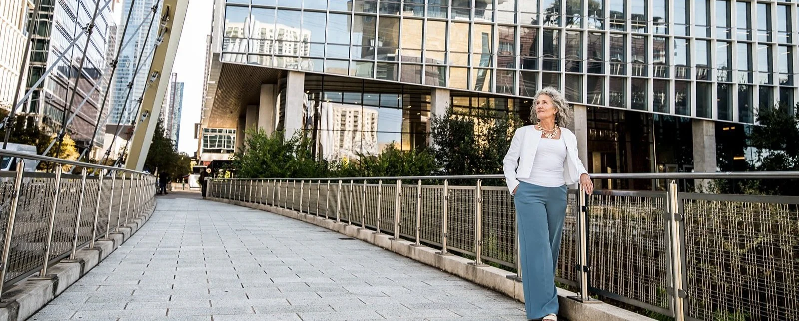 Ellen Lindsey, executive coach, in downtown Austin with gray curly hair walking along a city sidewalk, dressed in a white top, white blazer, and blue pants. She is looking to her right and carrying a small black bag.