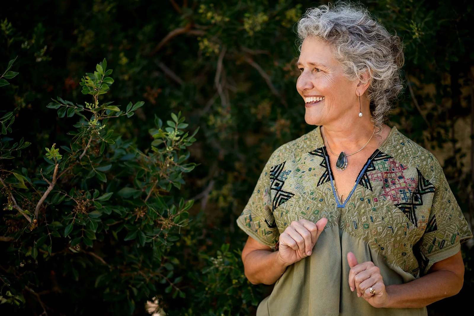 An elderly woman with curly gray hair smiling and looking to her right while jogging outdoors among green bushes, wearing a patterned blouse and jewelry.