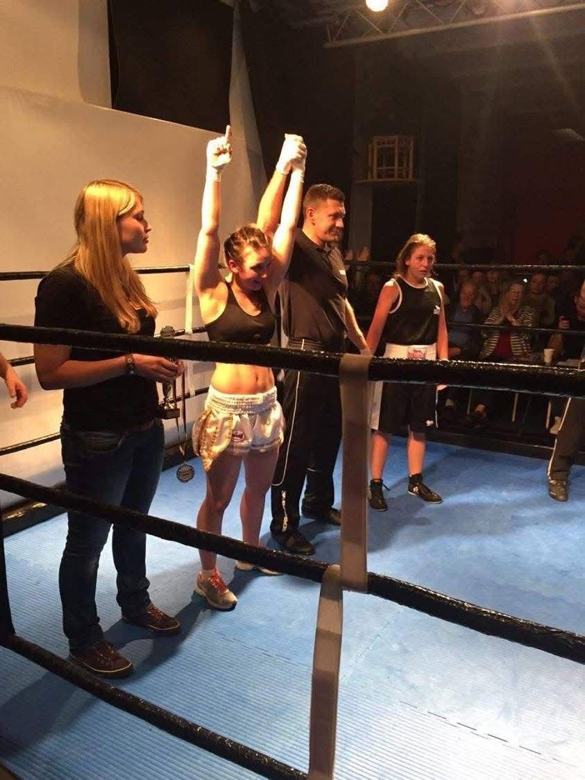Eine Boxeratcheszene in einem Ring mit vier jungen Frauen, eine befindet sich in Boxerpose mit erhobenen Armen, die anderen stehen neben ihr, im Hintergrund schauen Menschen zu.