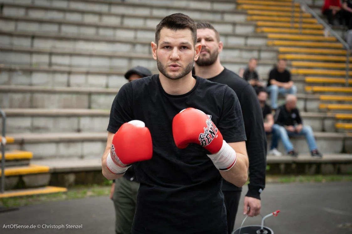 Mann mit schwarzen T-Shirt und roten Boxhandschuhen, der in Kämpferhaltung posiert, im Hintergrund einige Zuschauer sitzend auf Stufen.